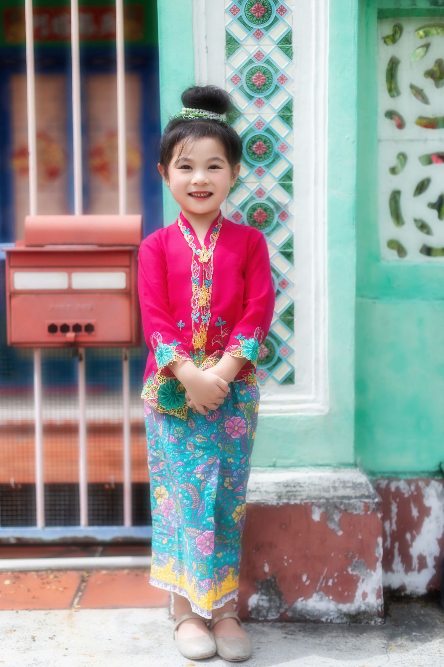 Photo of a Young girl wearing Nyonya Kebaya smiles in front of decorative Peranakan patterned wall, at Joo Chiat along Koon Seng road.