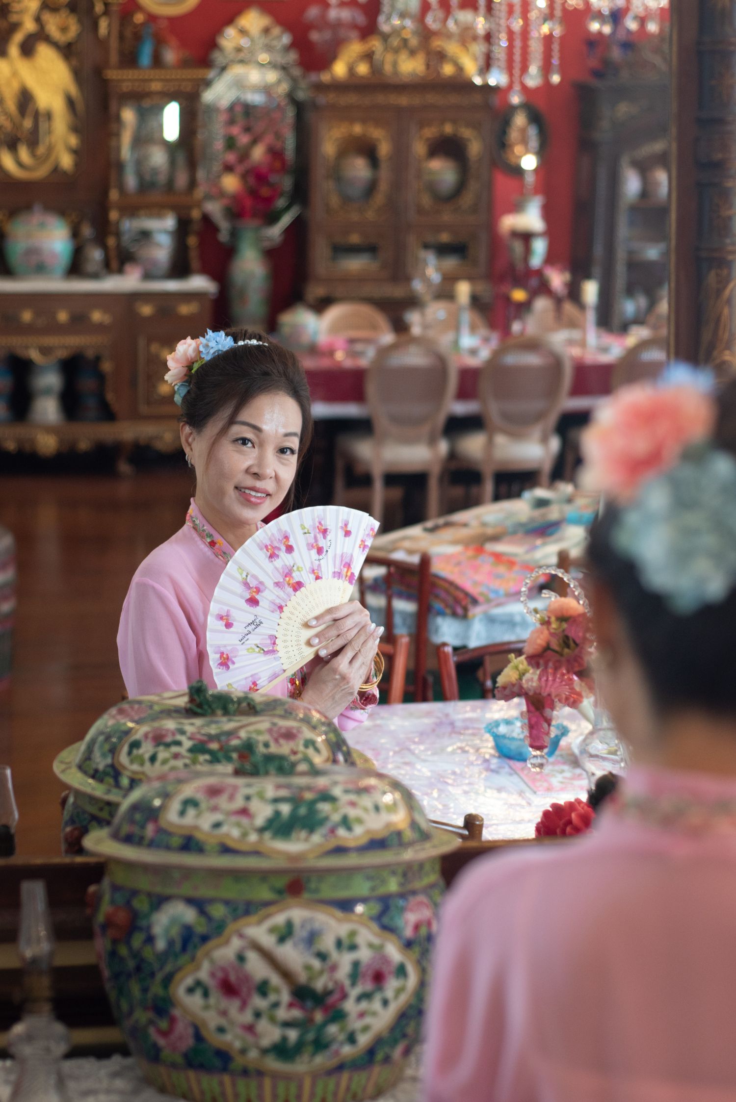 Traditional Asian decor fills an ornate dining room as someone in pink holds a decorative fan at an elegant table setting.