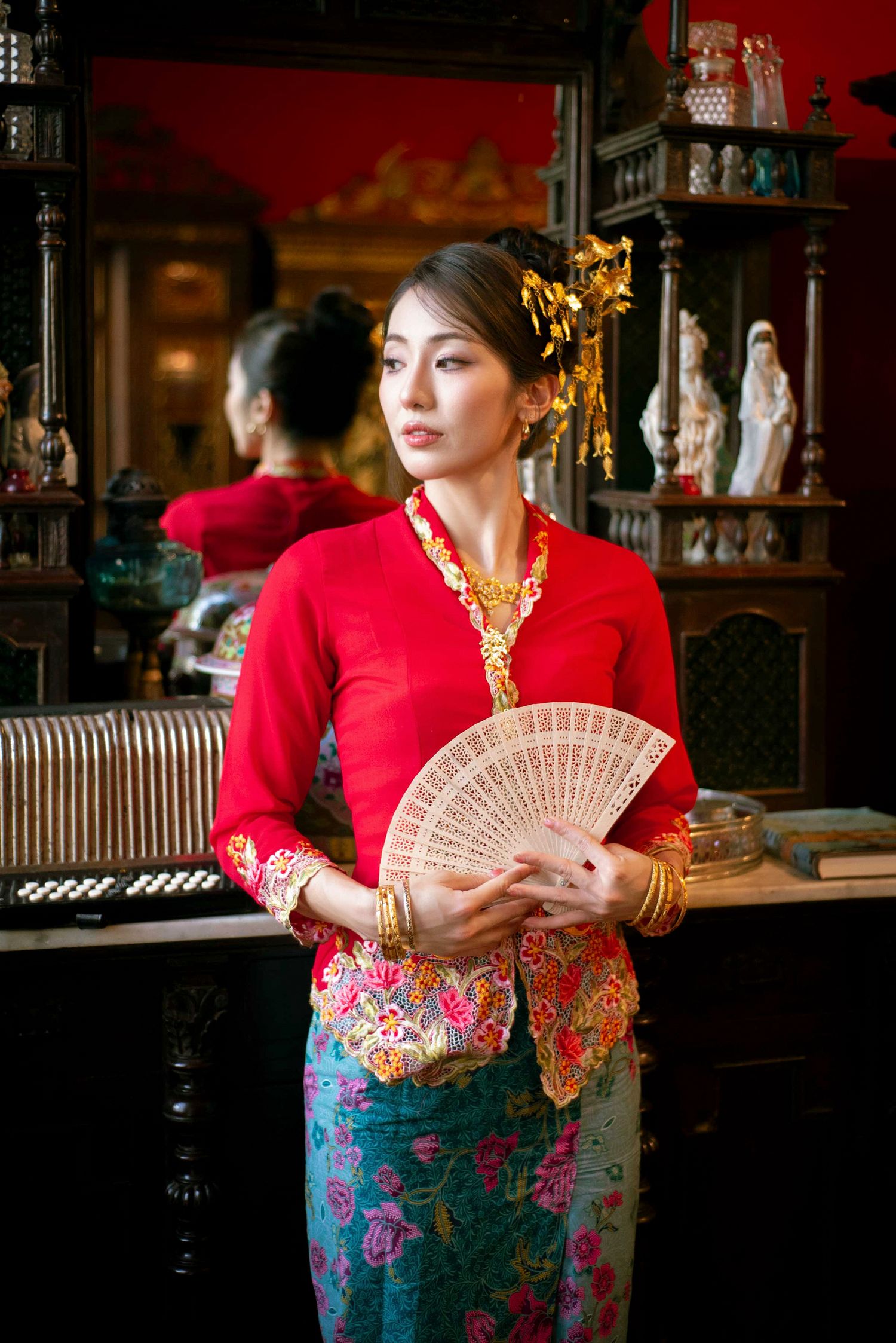 Woman in traditional red kebaya and batik skirt holds wooden fan while standing in ornate interior setting.