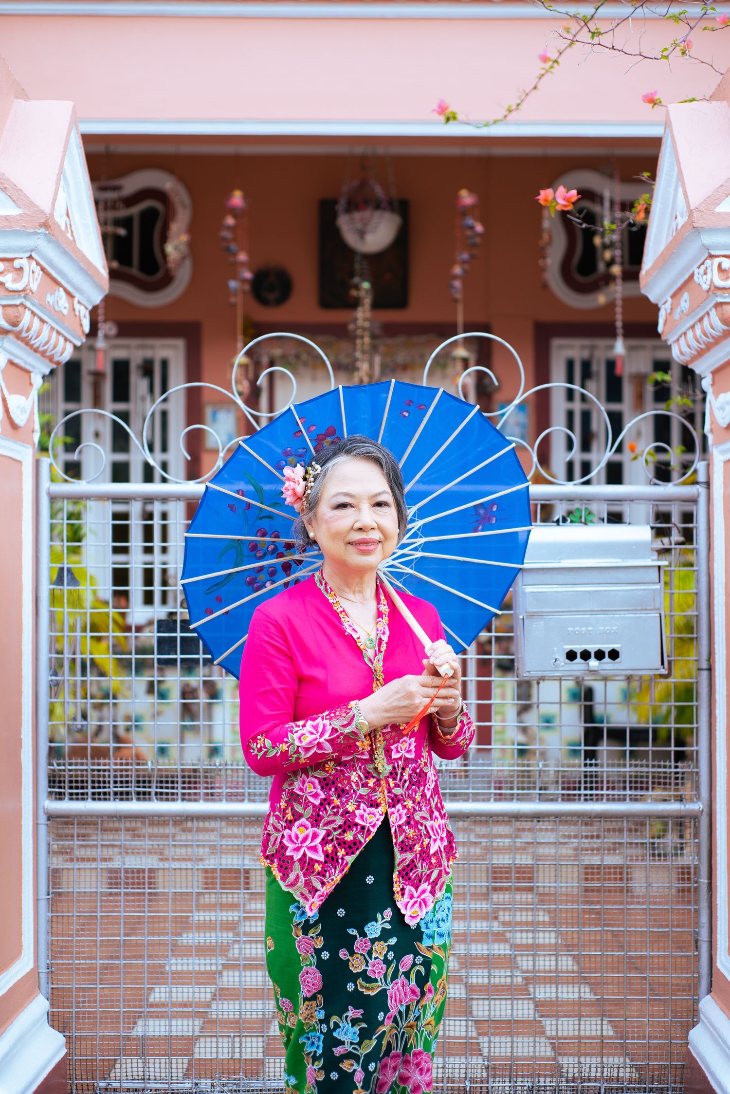 Traditional Southeast Asian woman holding blue parasol wearing pink kebaya and floral sarong standing in front of decorative building.