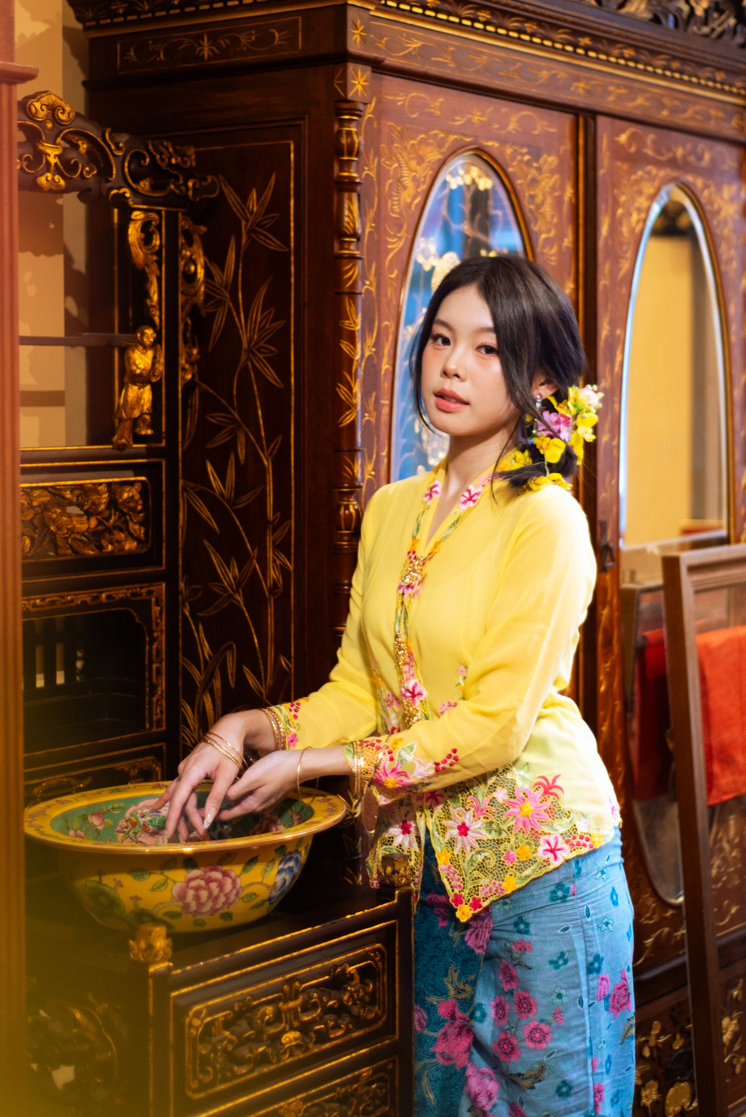 Woman in traditional Peranakan Nyonya kebaya outfit with yellow top and blue floral sarong poses against ornate backdrop.