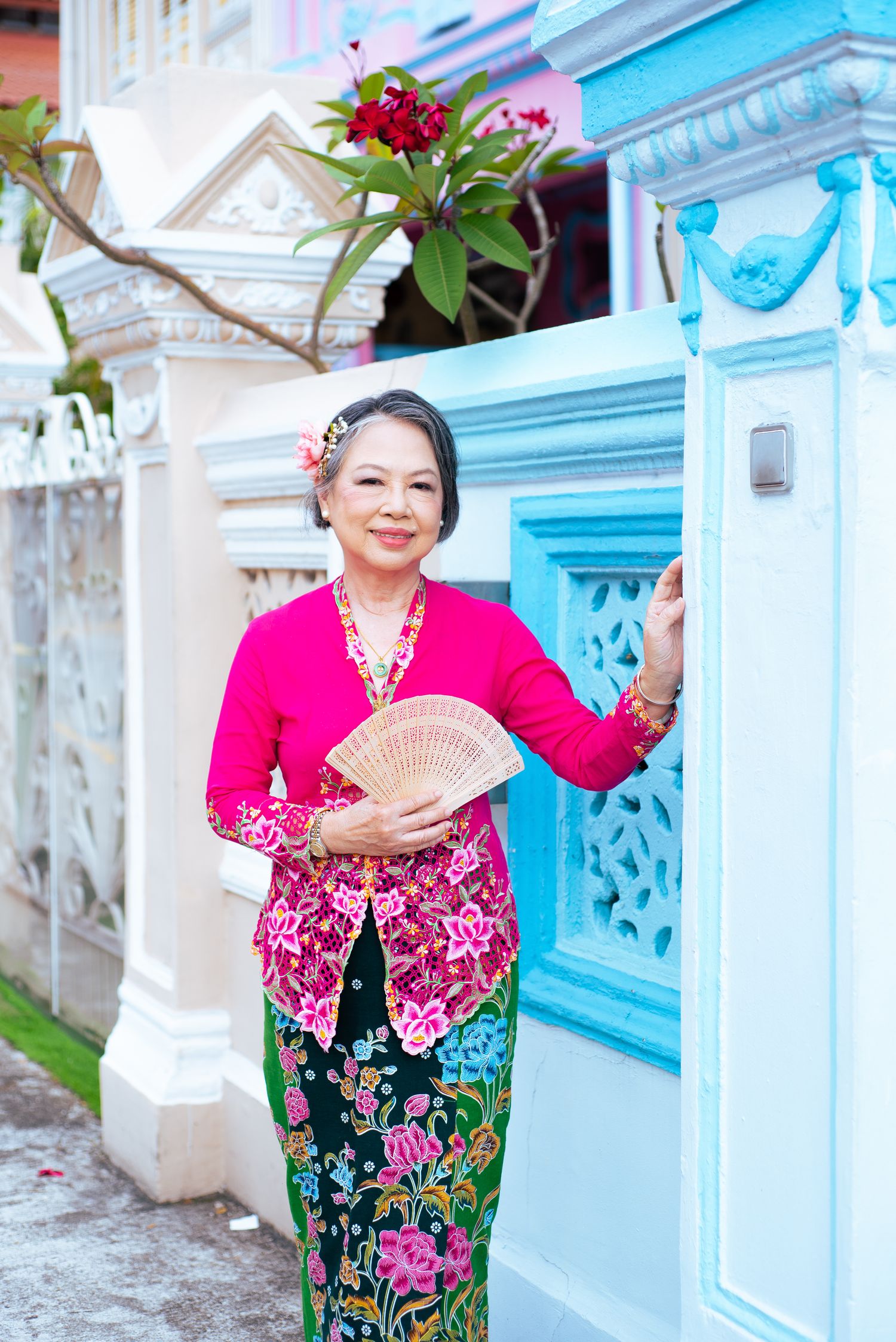 Traditional Peranakan kebaya fashion in pink and floral print worn with handheld fan against blue and white heritage wall.