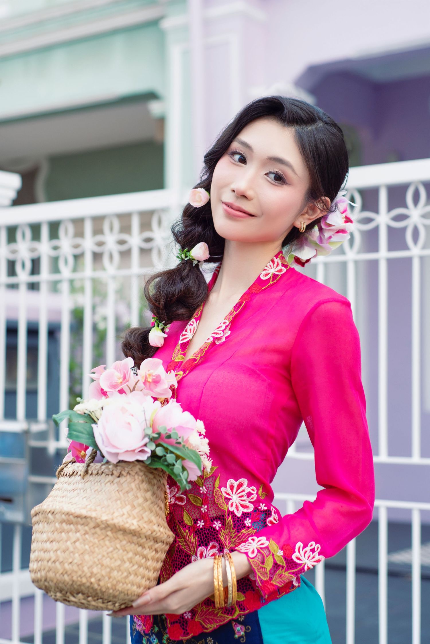 A person in a bright pink traditional Asian blouse holds a woven basket filled with pink roses and wears floral hair accessories.