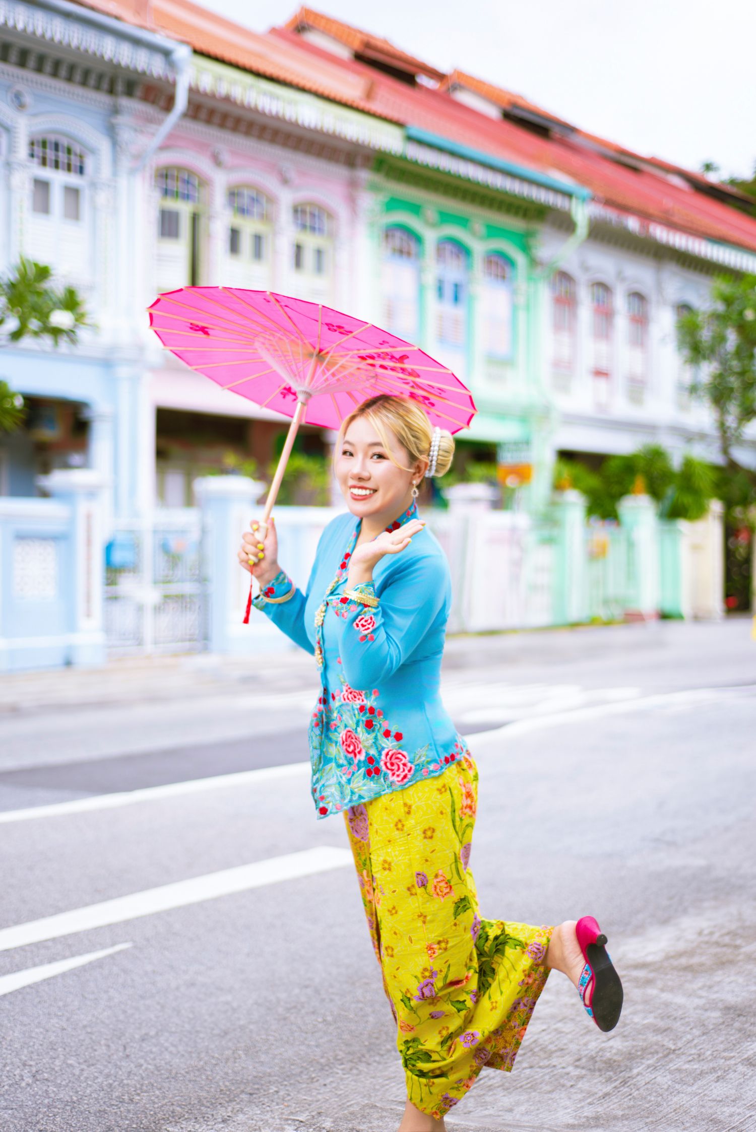 Colorful traditional Peranakan fashion with pink parasol against pastel shophouses in Singapore.