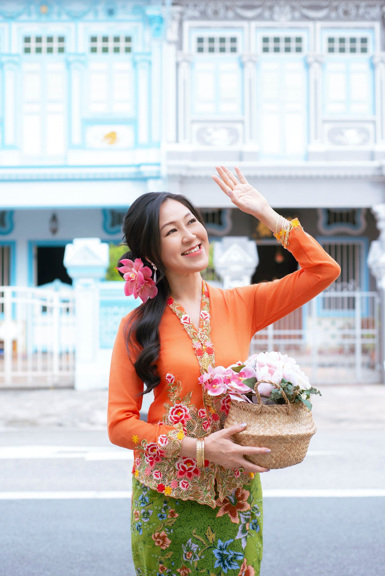 Woman in bright orange top and floral green sarong carries basket of flowers while waving on city street.