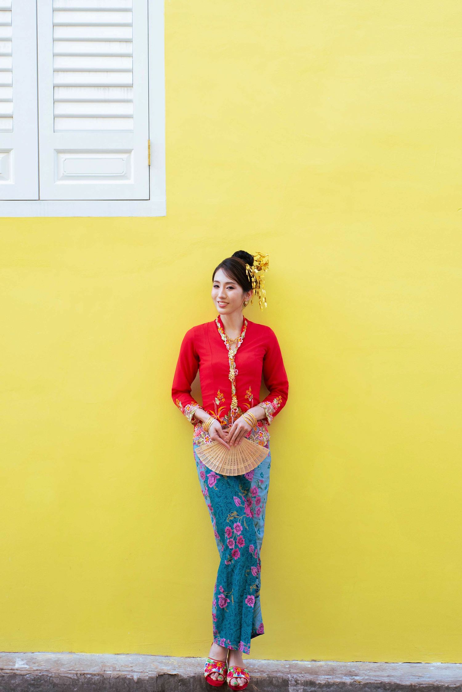 Woman in traditional Peranakan Nyonya outfit with red kebaya top and batik sarong against bright yellow wall.