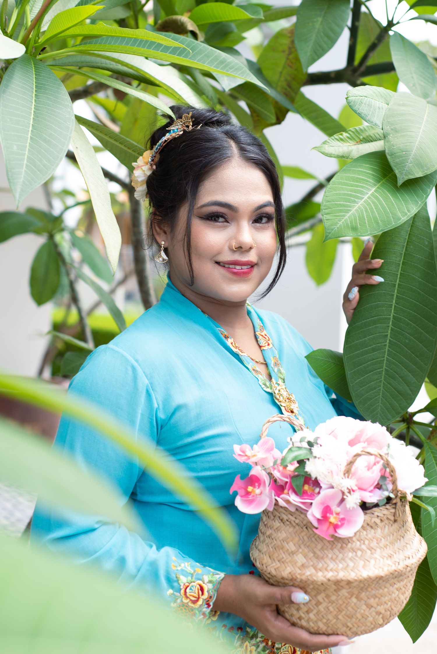 Person in turquoise traditional dress holds decorative basket of pink flowers among tropical green foliage.