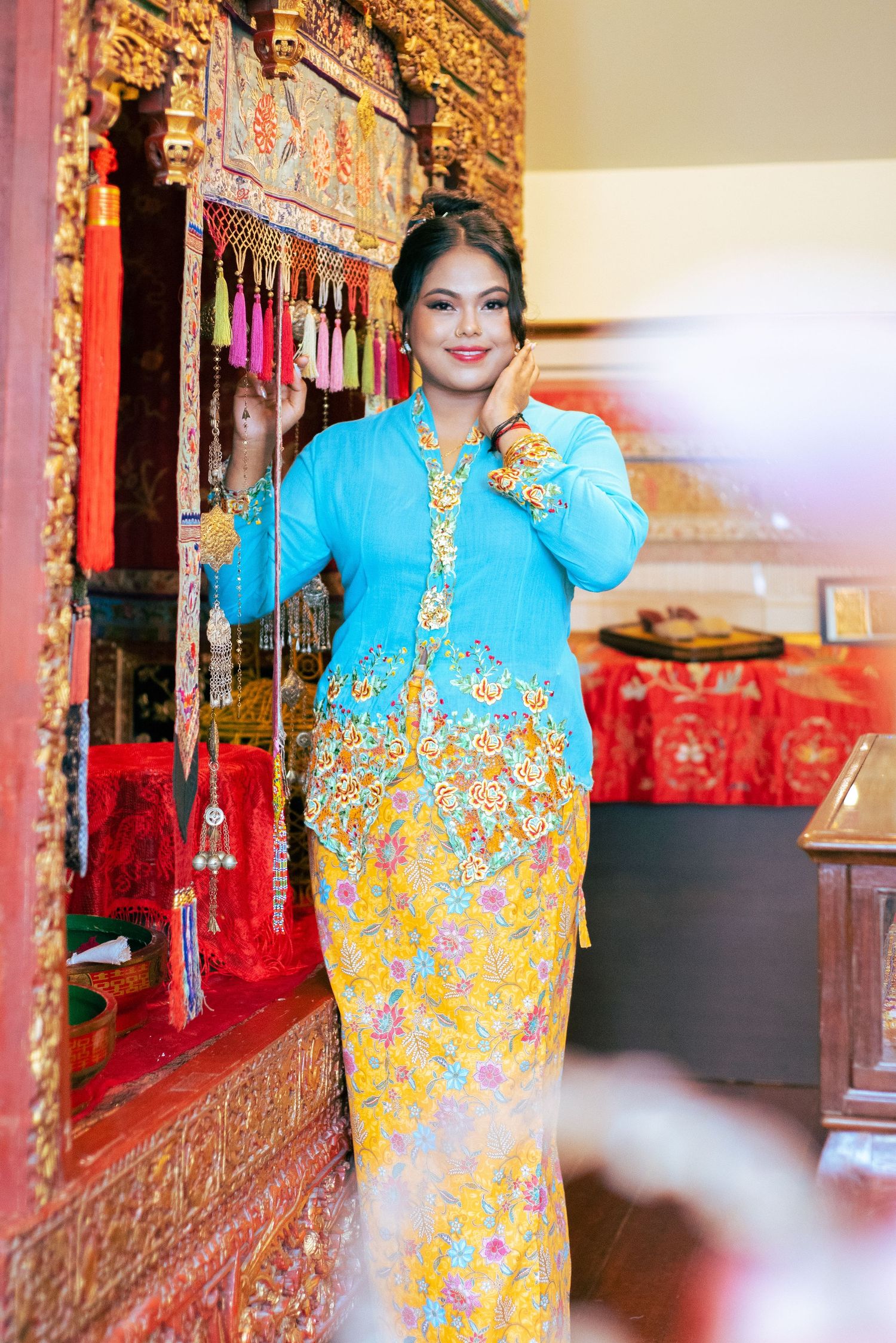 Someone wearing a traditional Balinese kebaya dress in turquoise and yellow with ornate floral patterns stands by a temple altar.