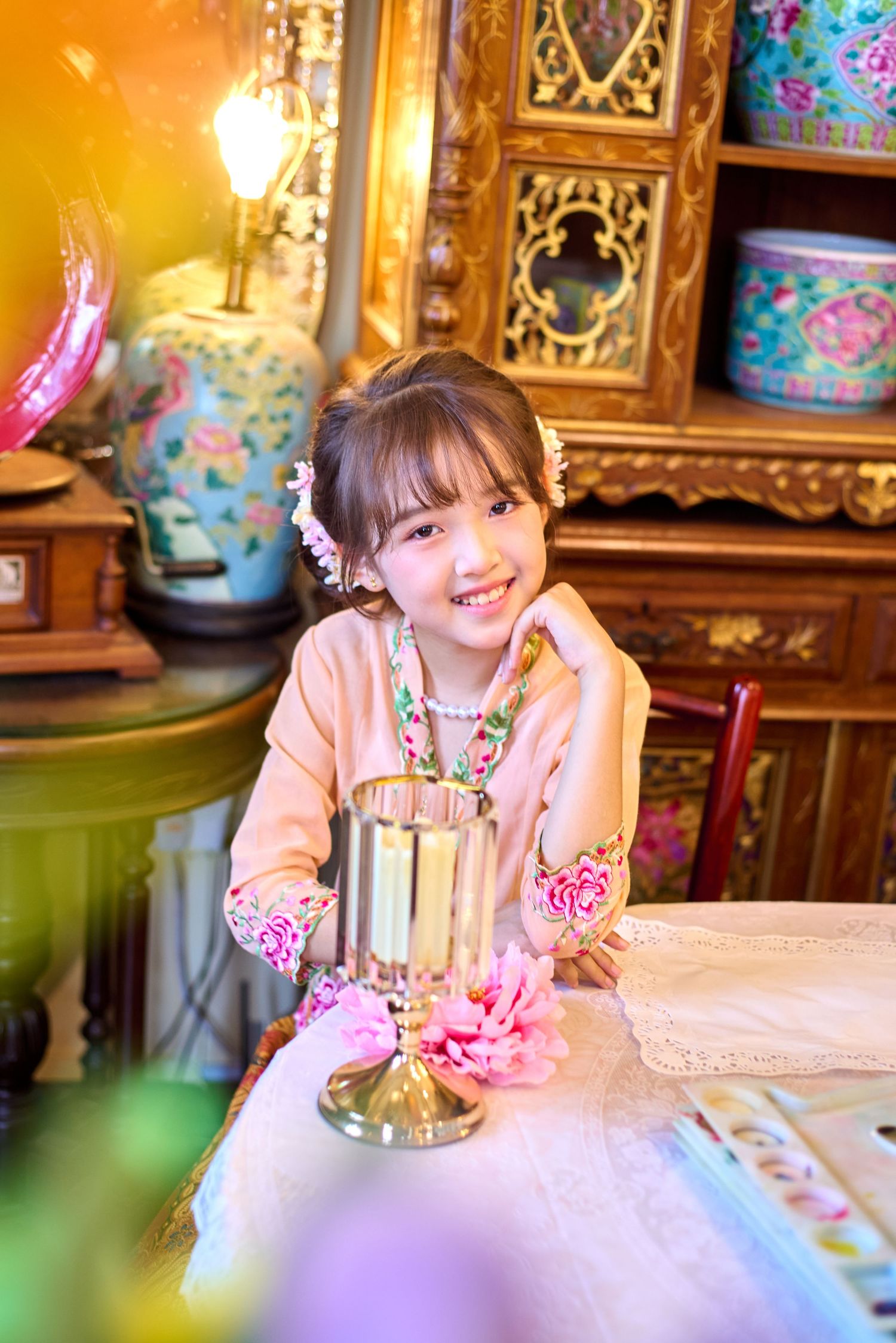 Young girl in pink floral traditional dress smiling at ornate table with golden candleholder and colorful ceramic vases.