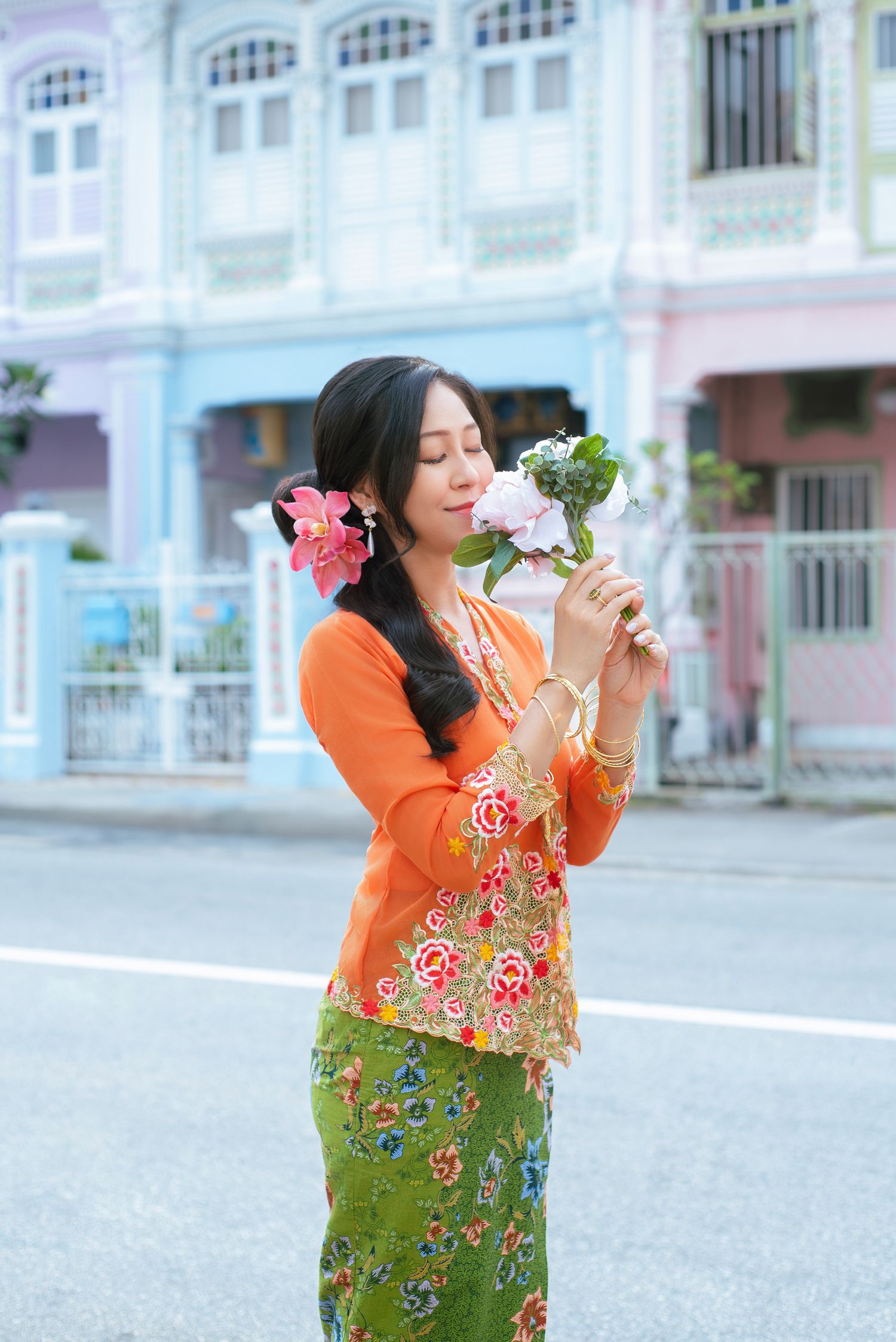 A person in an orange top and floral green skirt poses with flowers in an outdoor urban setting.
