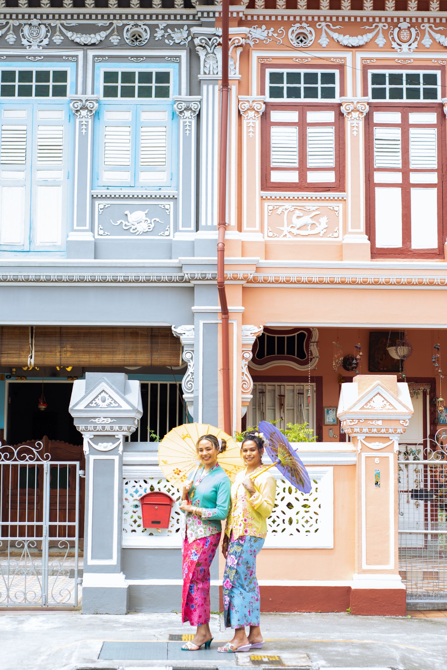 People in traditional clothing walking outside colorful vintage shophouses with ornate architectural details in Singapore.