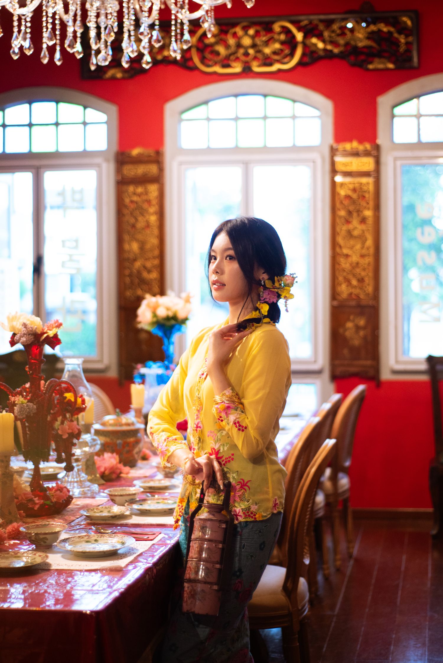 A woman in a yellow traditional floral blouse stands in an elegant red dining room with ornate windows and crystal chandeliers.