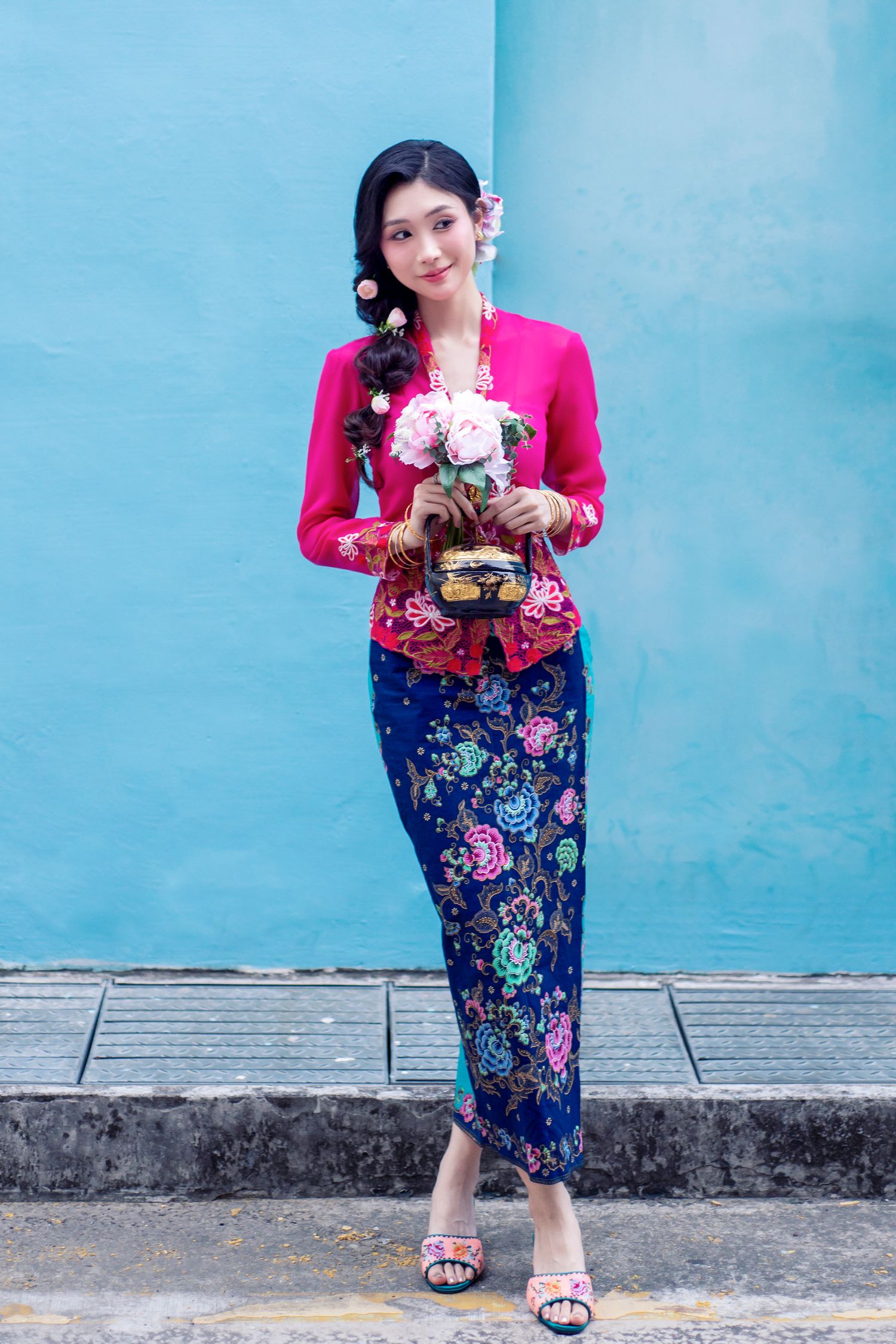 Traditional Peranakan outfit featuring a fuchsia kebaya top and blue floral sarong against a turquoise wall background.