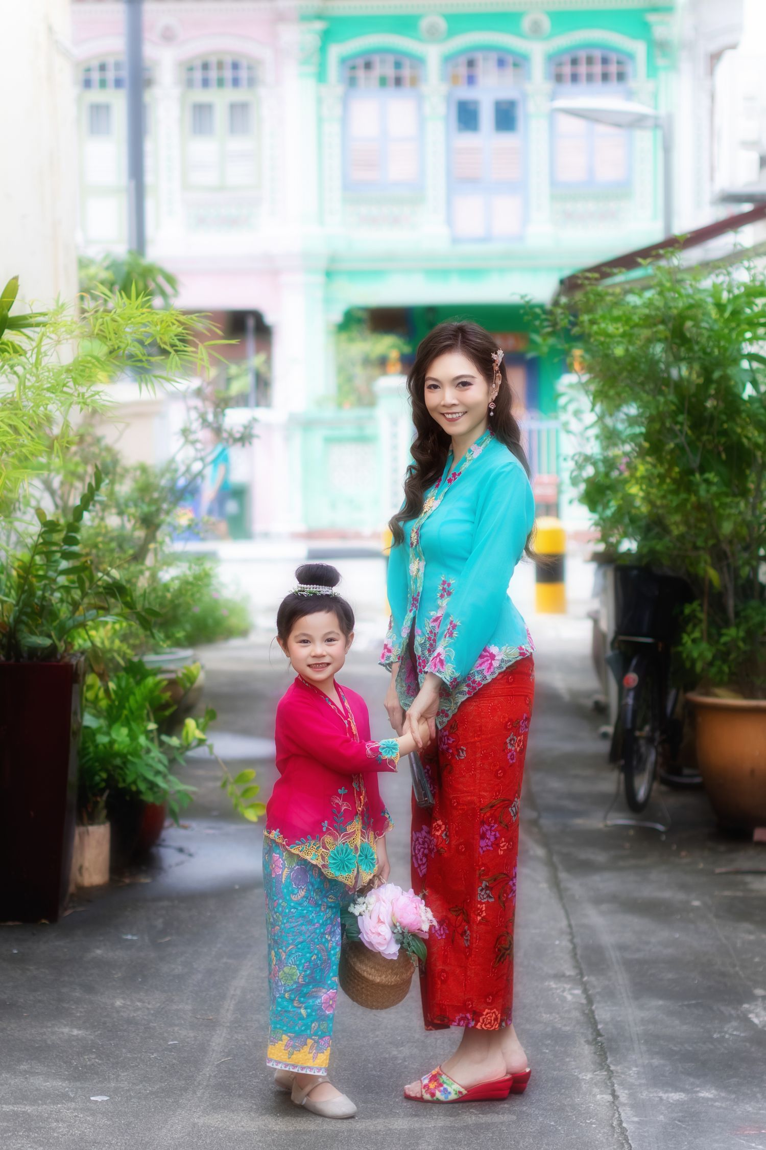Mother and daughter in traditional Nyonya Kebaya walking together carrying a basket of flowers, in front of iconic Peranakan houses.