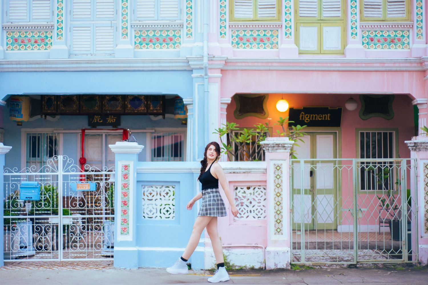 A person in a skirt poses in front of colorful pastel shophouses with ornate architectural details in Singapore.