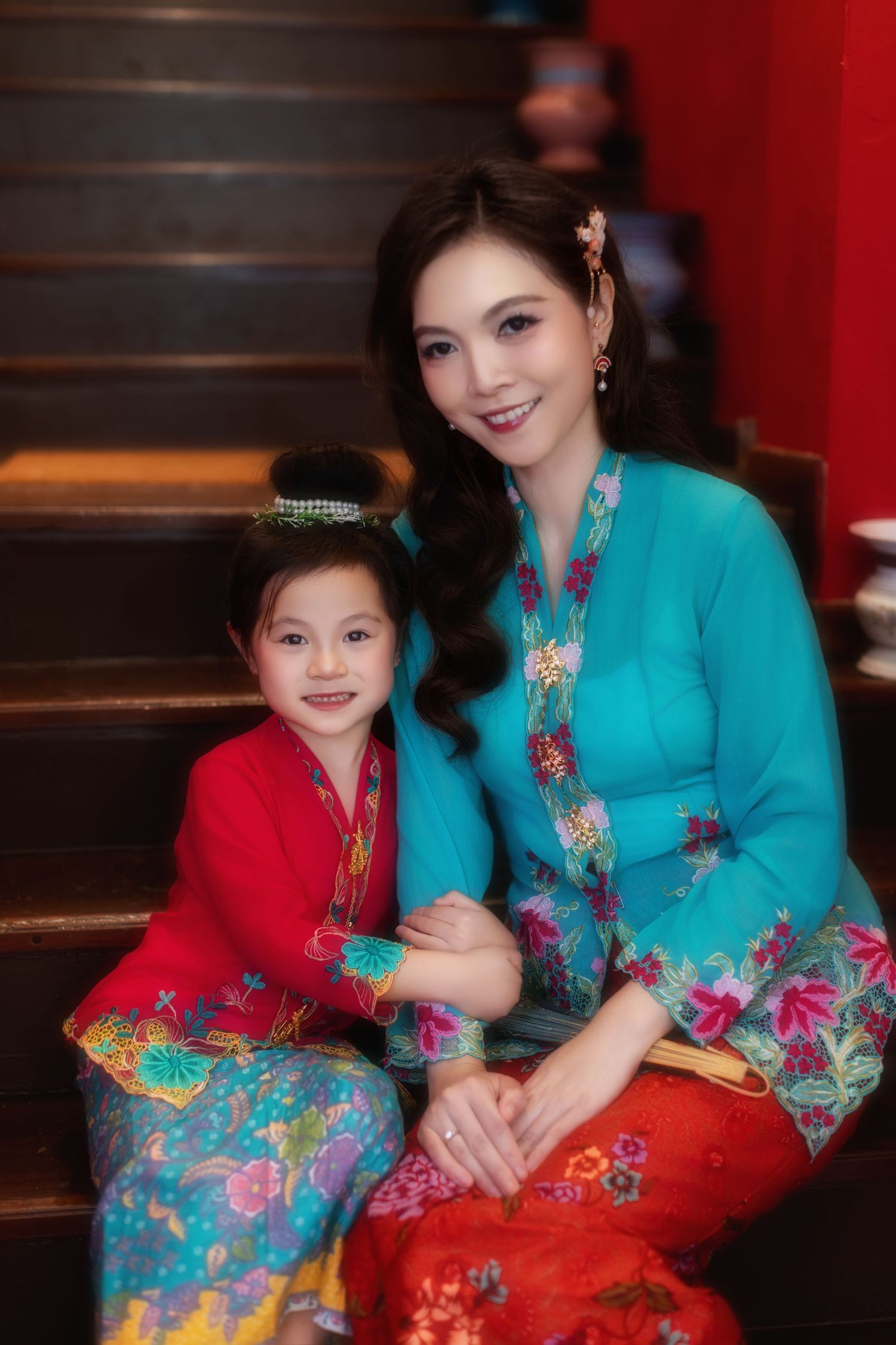 Photo of a Mother and daughter in traditional Nyonya Kebaya sitting on red-walled stairs with decorative vases.