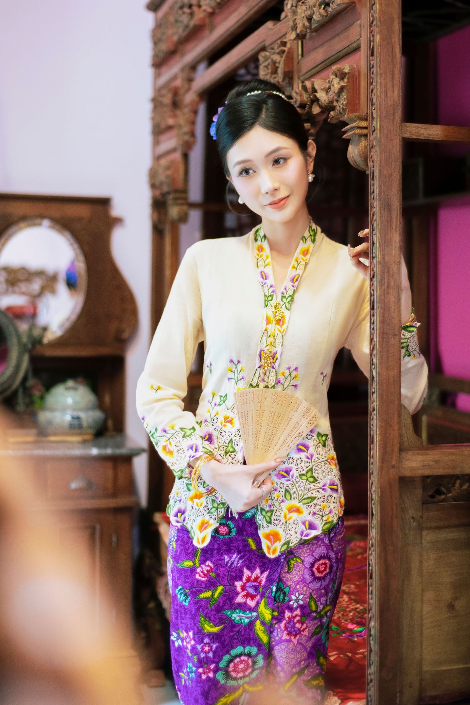 Traditional Southeast Asian outfit featuring a cream kebaya top with floral embroidery and bright purple batik sarong skirt.
