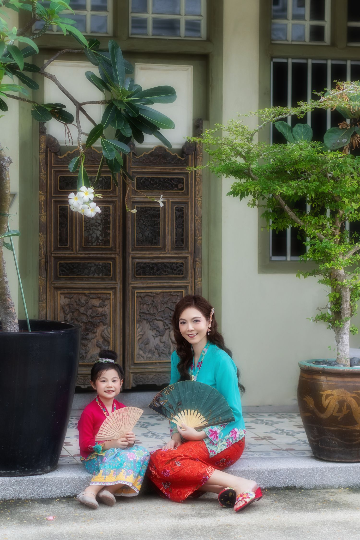 Photoshoot of mother and daughter in traditional Nyonya Kebaya sitting down against ornate wooden door Pintu pagar.