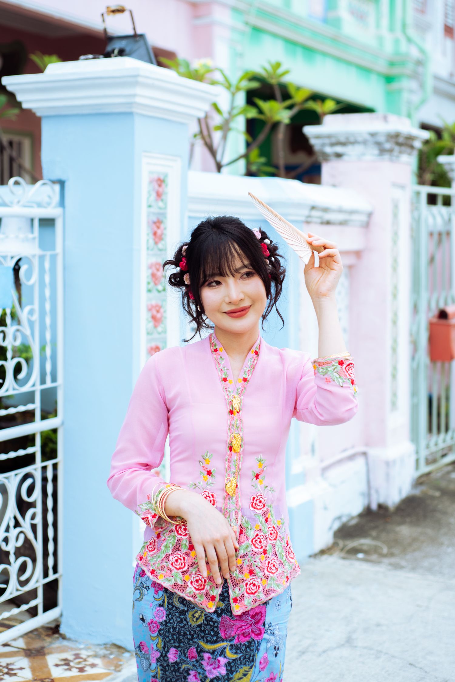 A woman wearing a pink floral blouse and patterned skirt poses next to white decorative pillars in an outdoor setting.