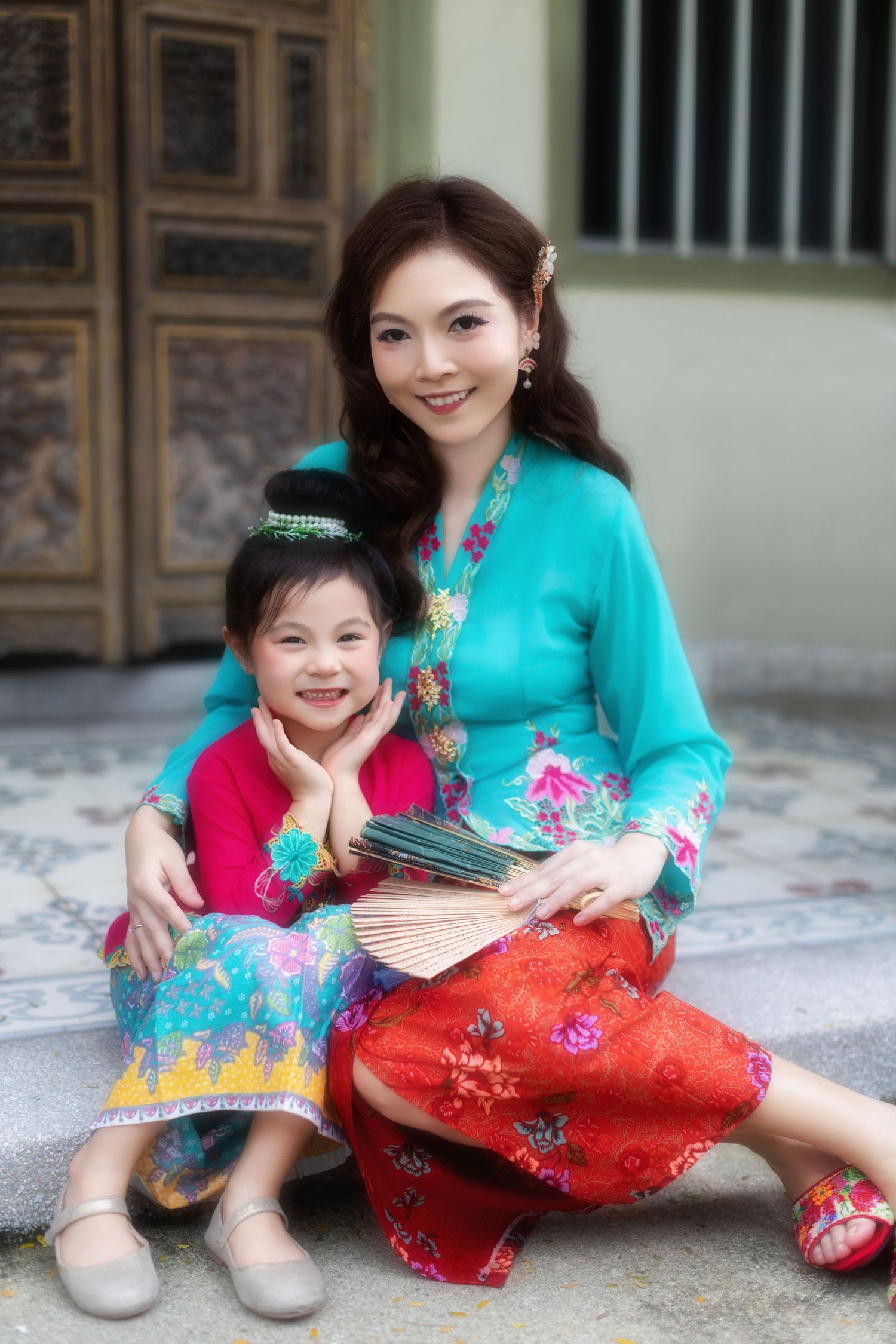 Portrait of a Mother and girl in traditional Nyonya Kebaya sitting down and smiling, in front of a Peranakan house.
