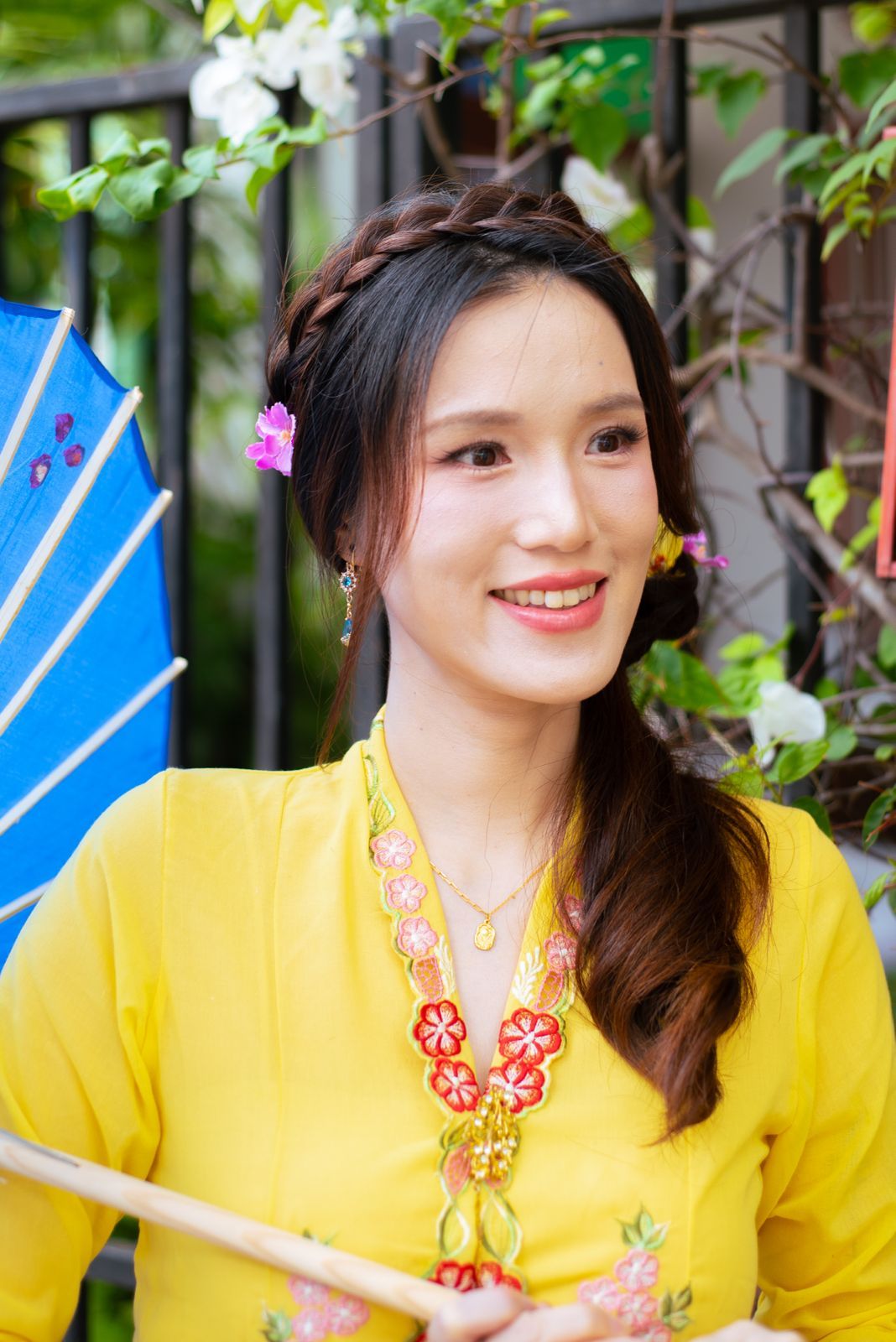 A figure in a bright yellow traditional dress with floral accessories smiles while holding a blue paper fan against garden foliage.