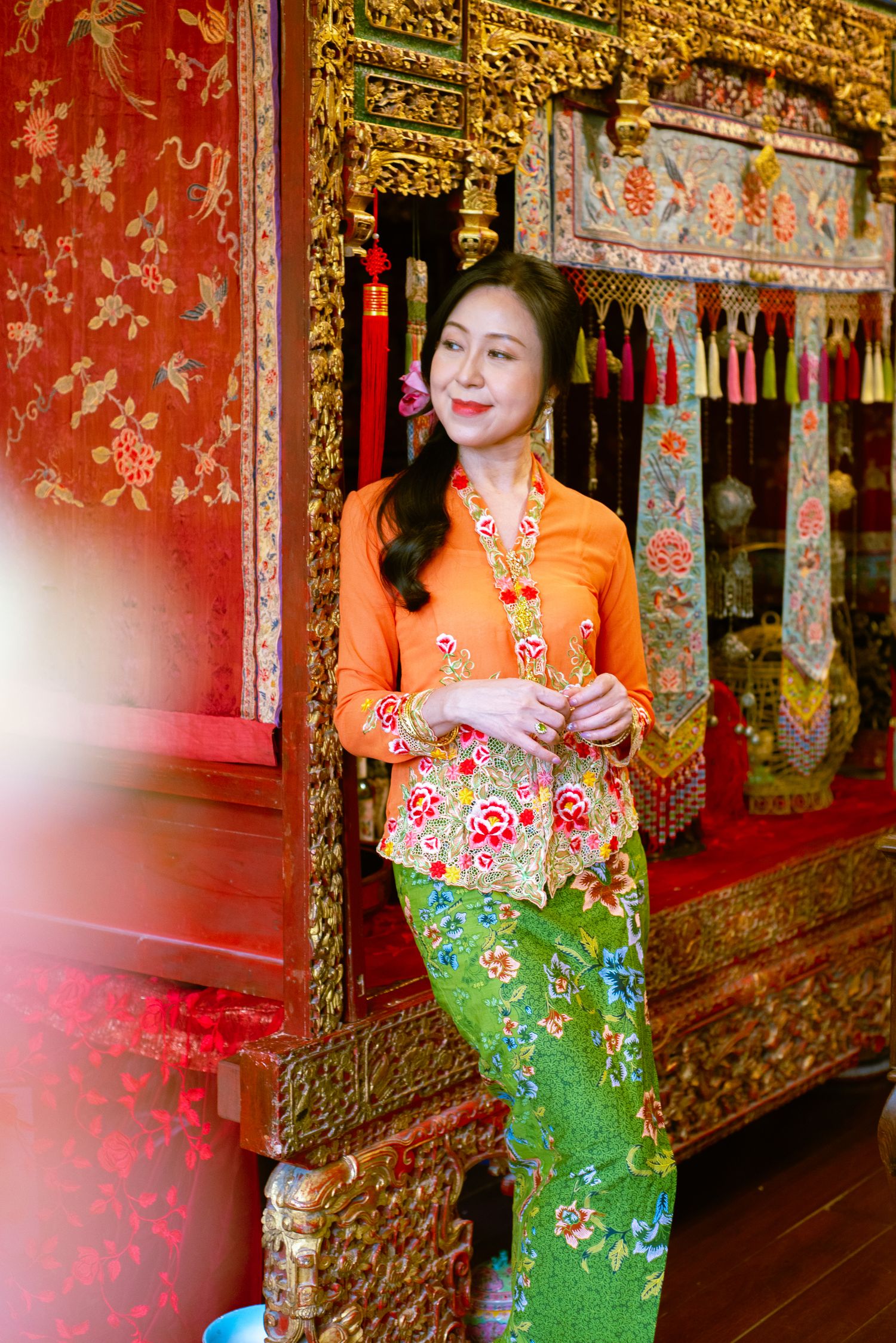 Traditional Southeast Asian clothing featuring an orange kebaya top and green batik skirt photographed at an ornate temple doorway.