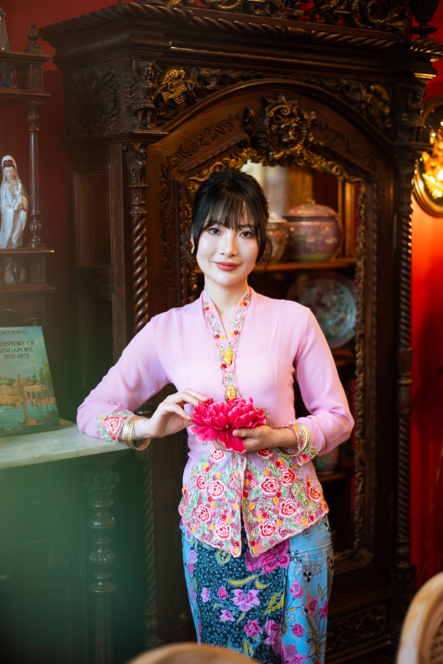 A person in a pink traditional kebaya top and floral sarong stands by an ornate wooden archway holding pink flowers.