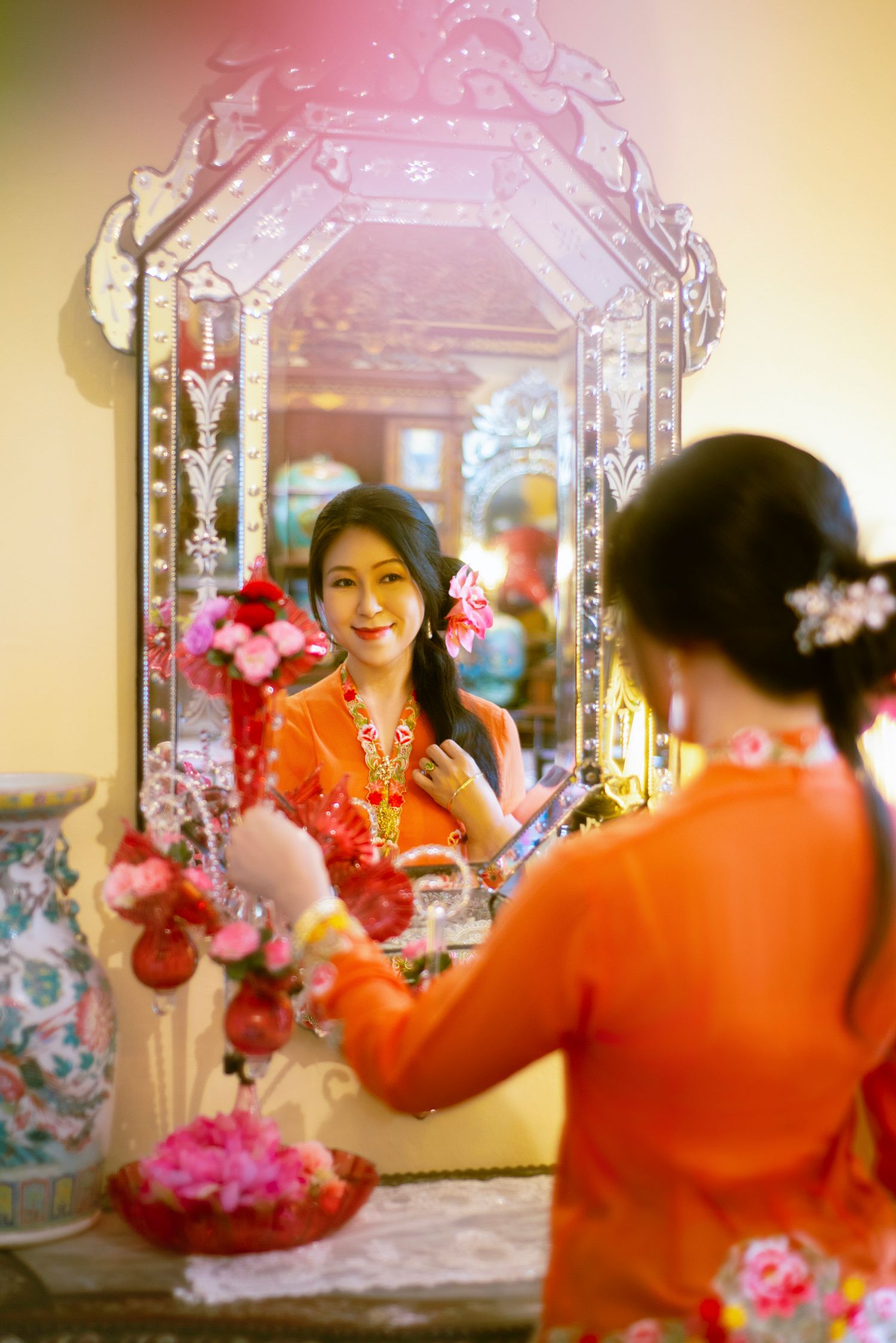 Person wearing orange traditional attire with pink flowers poses in front of an ornate decorative mirror.