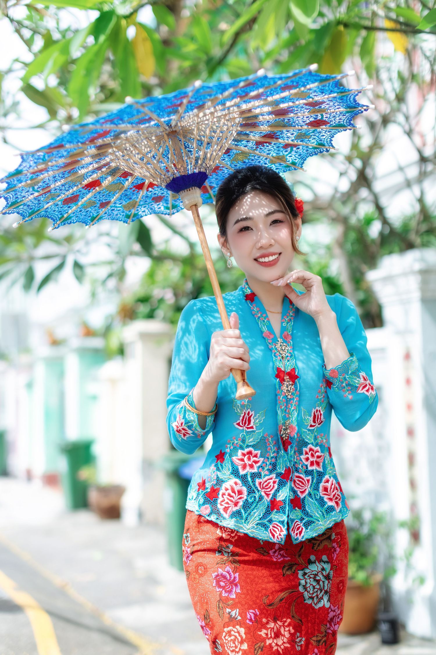 Three photos show someone in traditional Asian attire with a turquoise top, red skirt and decorative blue parasol against greenery.