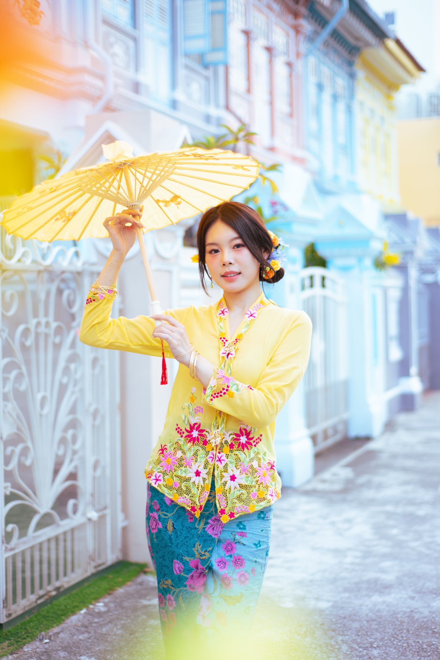 Person in traditional Asian attire with yellow cardigan and floral print holds parasol against colorful building backdrop.