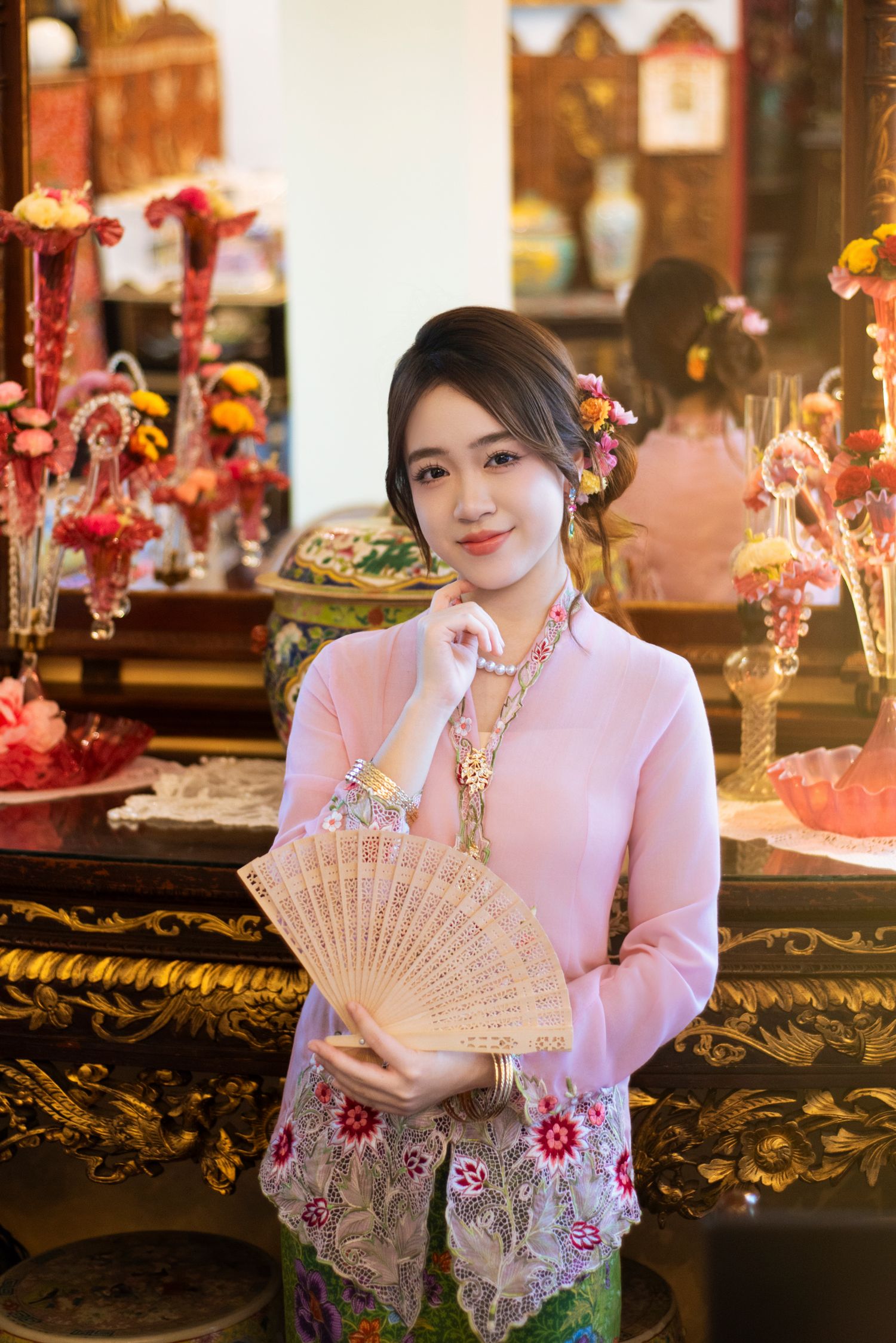Traditional Asian outfit with pink floral kebaya top and decorative fan in ornate interior setting.