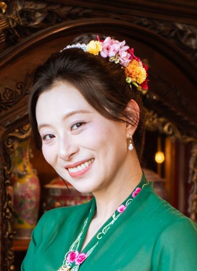 Person wearing a bright green kimono decorated with floral accessories and pink flower hair ornaments smiles warmly.