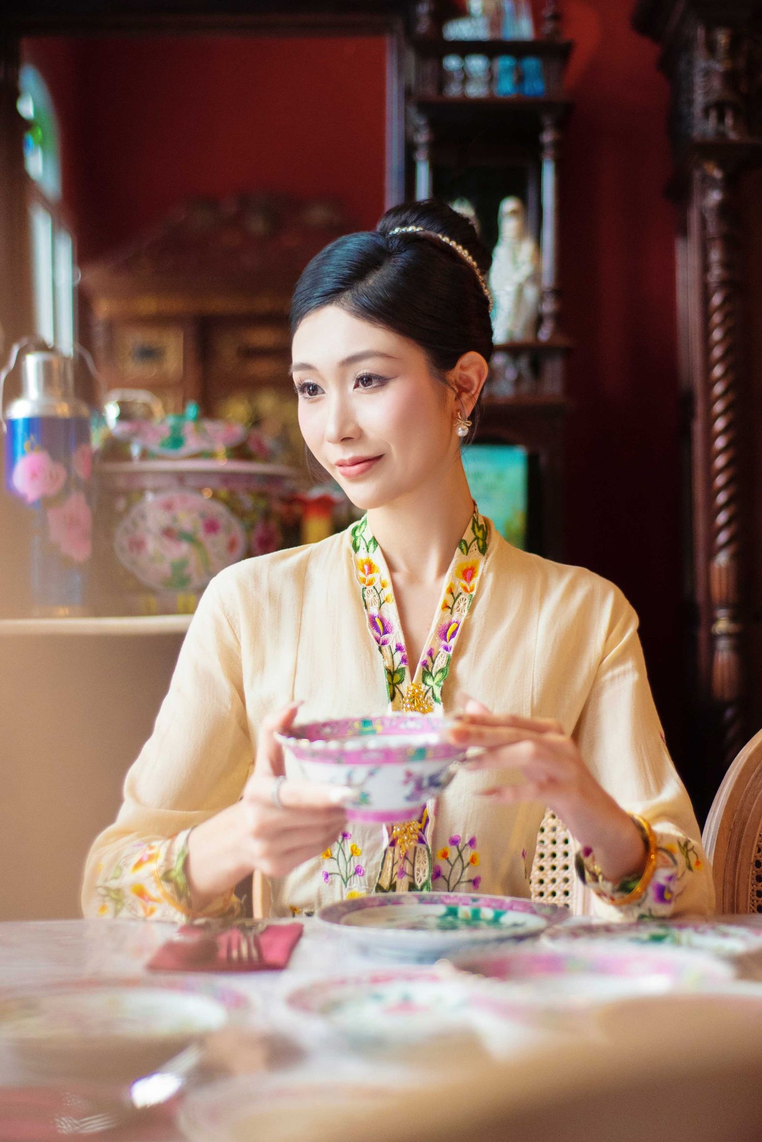 Woman in traditional Asian attire enjoys afternoon tea service with floral porcelain at an ornate restaurant.
