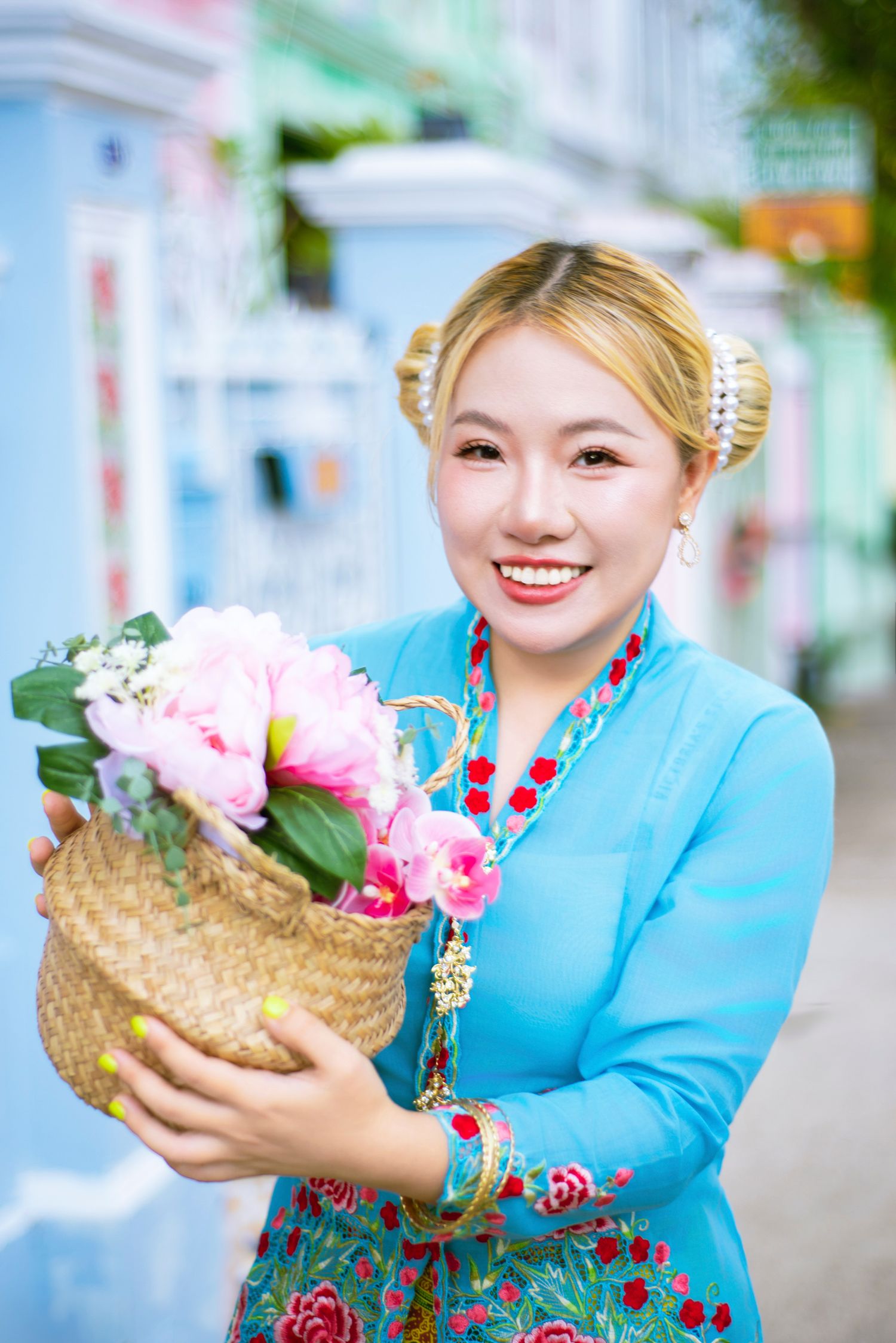 A person in a turquoise traditional dress with red embroidery holds a woven basket containing pink flowers.