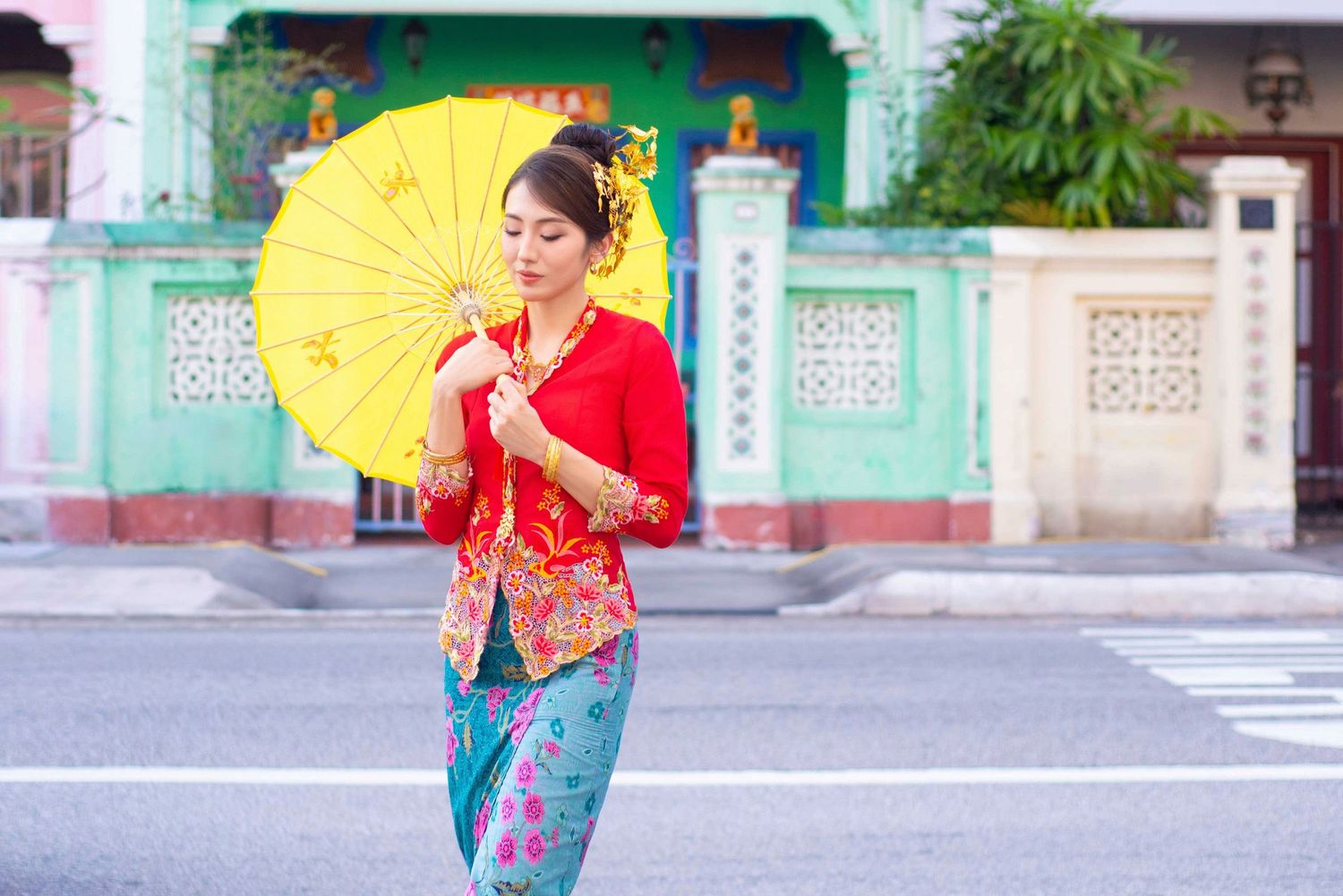 Traditional Asian fashion meets modern style with bright red top, jeans, and yellow parasol against turquoise building exterior.