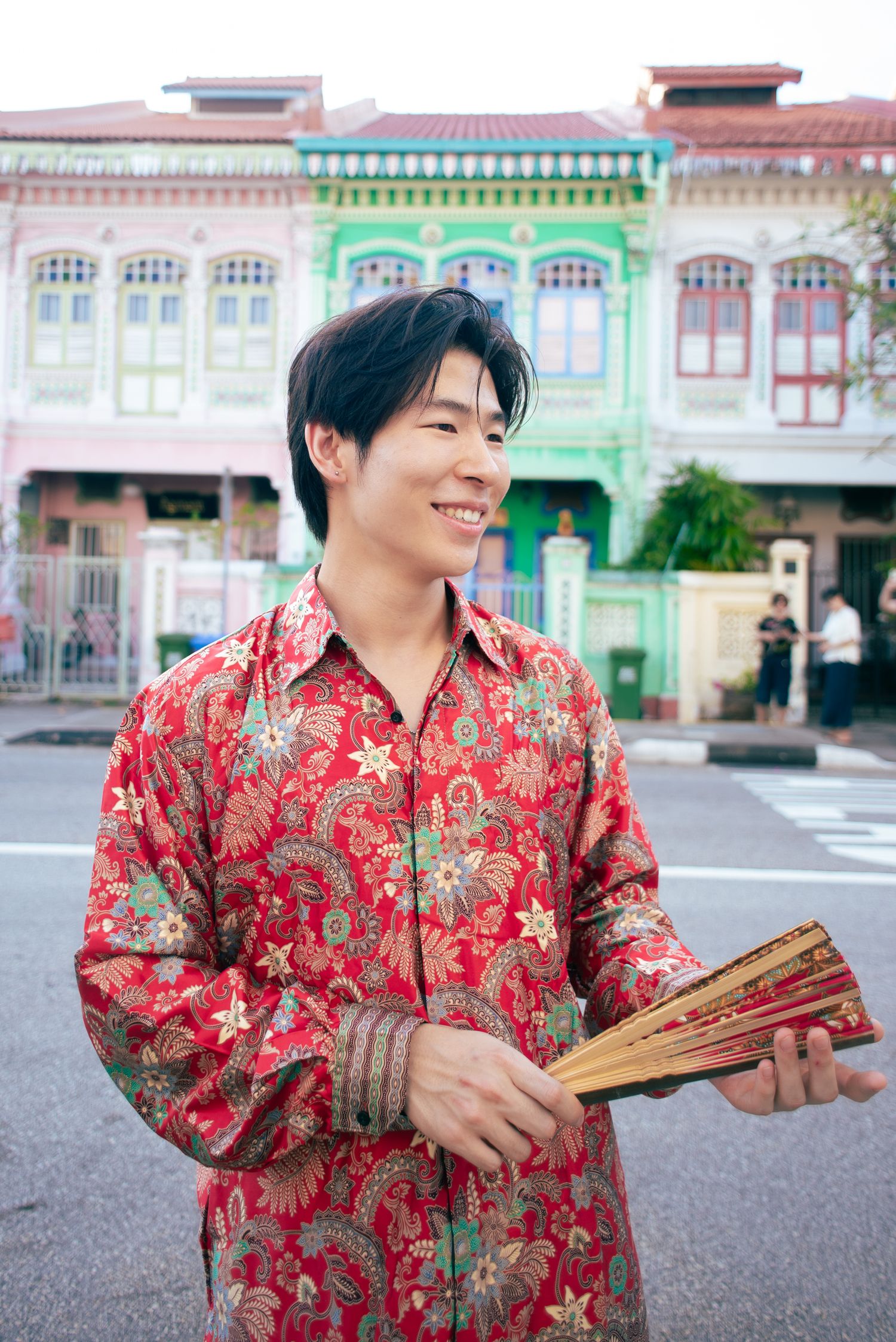 A person wearing a red floral traditional Chinese shirt holds a decorative fan in front of colorful heritage shophouses.