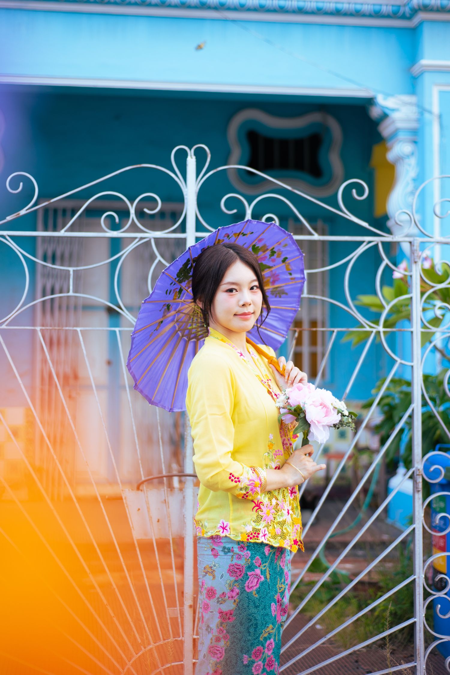 A person in traditional Burmese attire holds a purple parasol against an ornate blue and white gate.