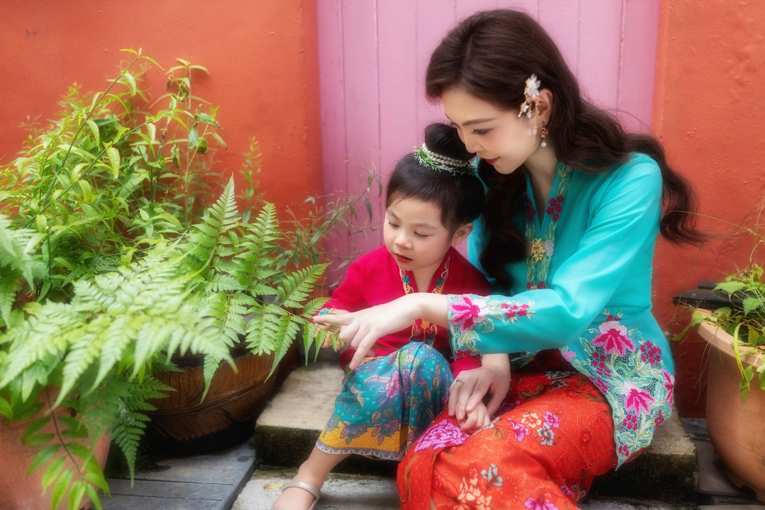 Photoshoot of a mother and daughter in traditional Nyonya Kebaya sitting down against a coral wall with pink door and garden details.