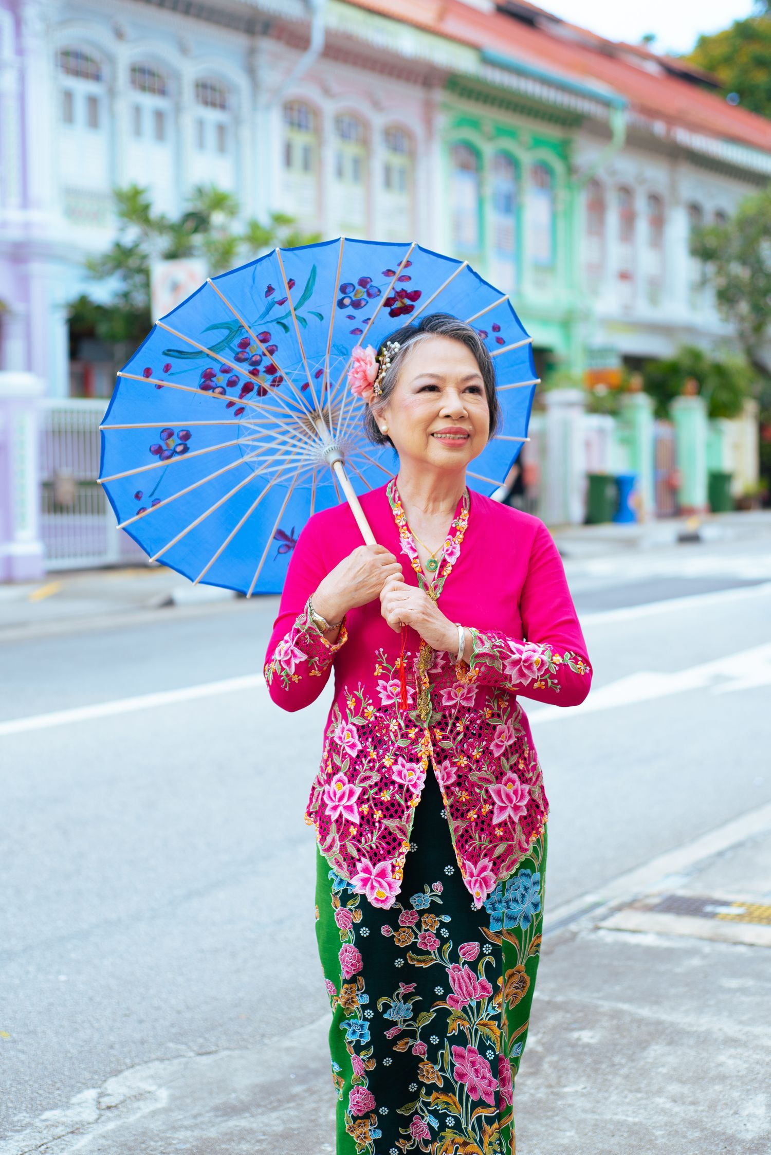 Traditional Peranakan nyonya outfit with bright pink kebaya and floral sarong walking with decorative blue parasol on street.
