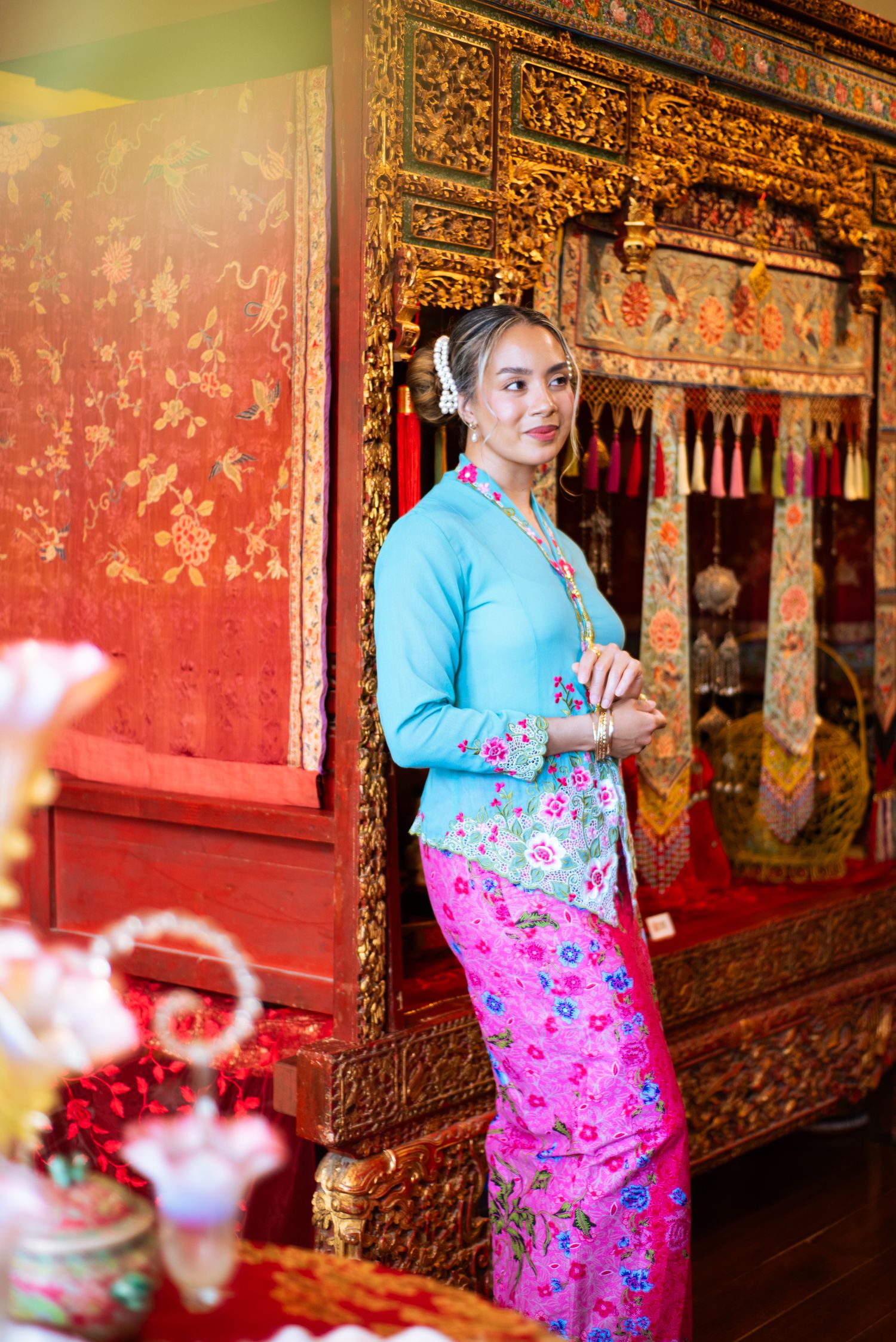 A person wearing a blue cardigan and pink floral dress stands in an ornate temple corridor with decorative red walls.