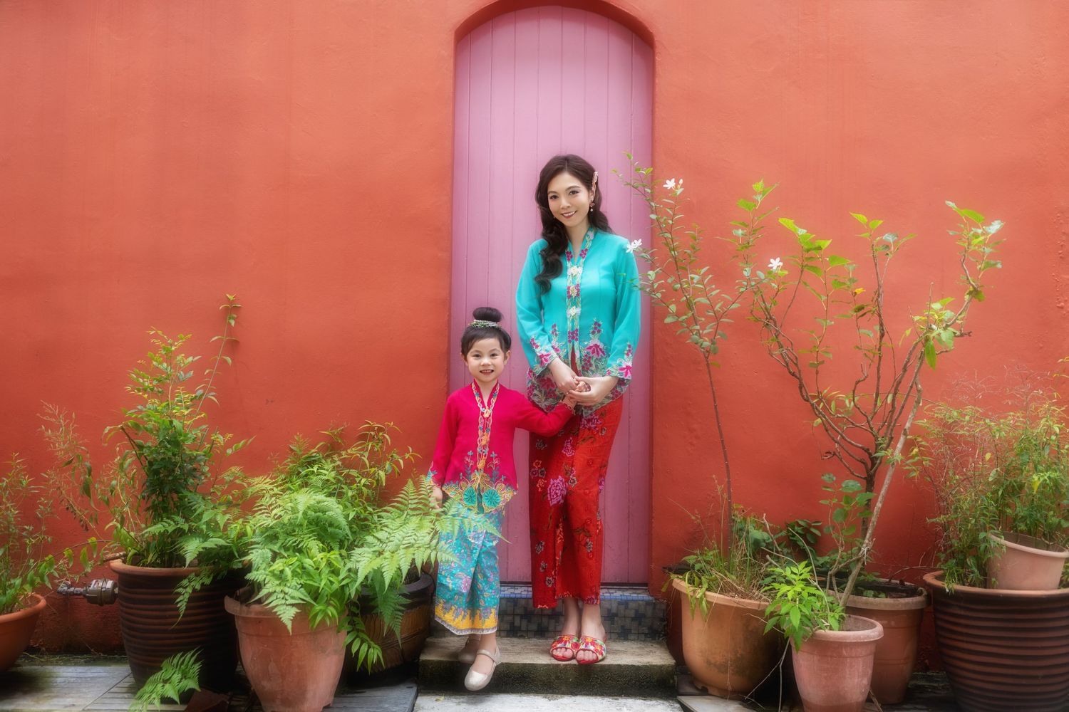 Candid fashion photo of Mother and daughter in traditional Nyonya Kebaya against a coral wall with pink door and garden details.