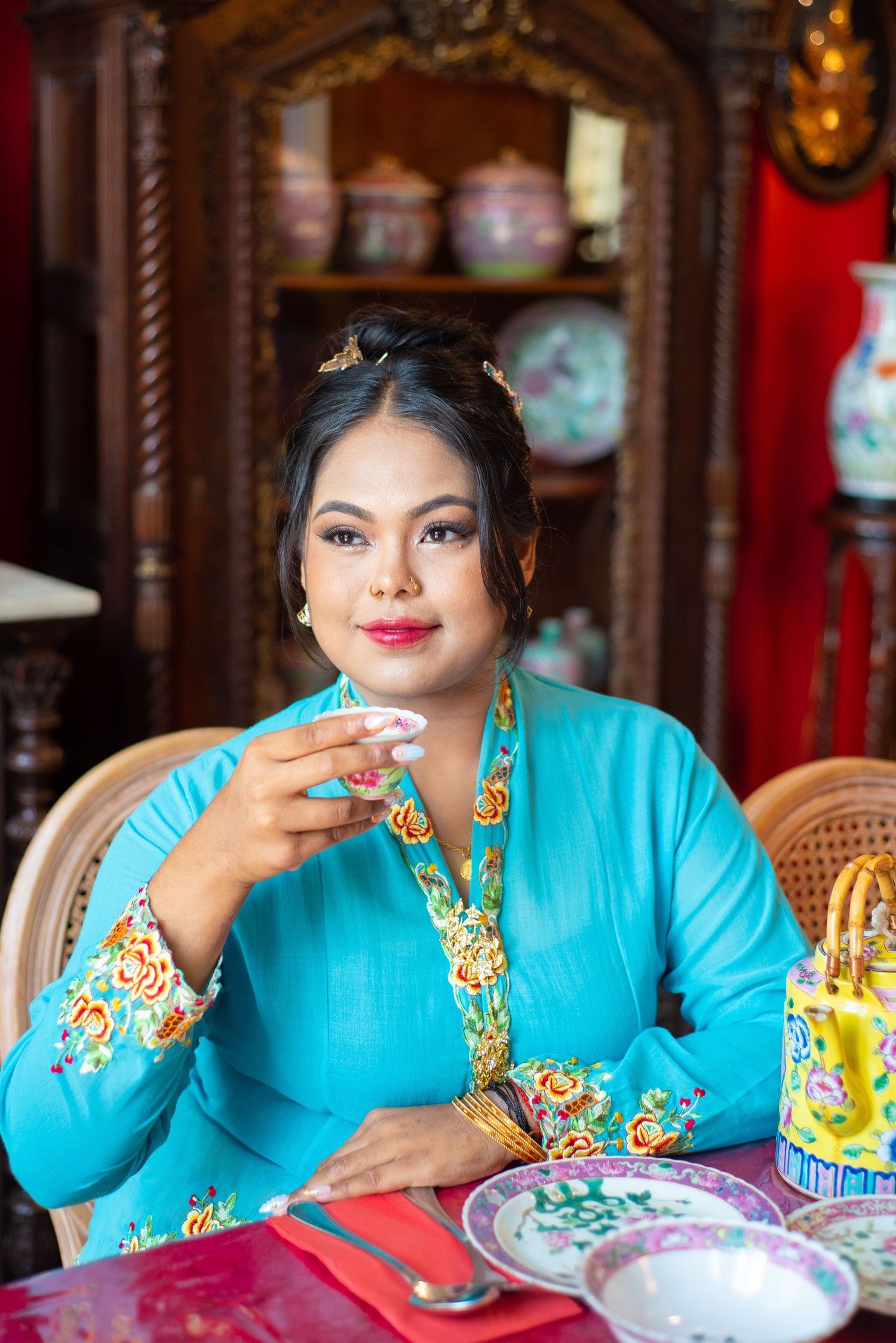 A person in a turquoise traditional dress with gold accents sits at a table in an ornate red-walled room.