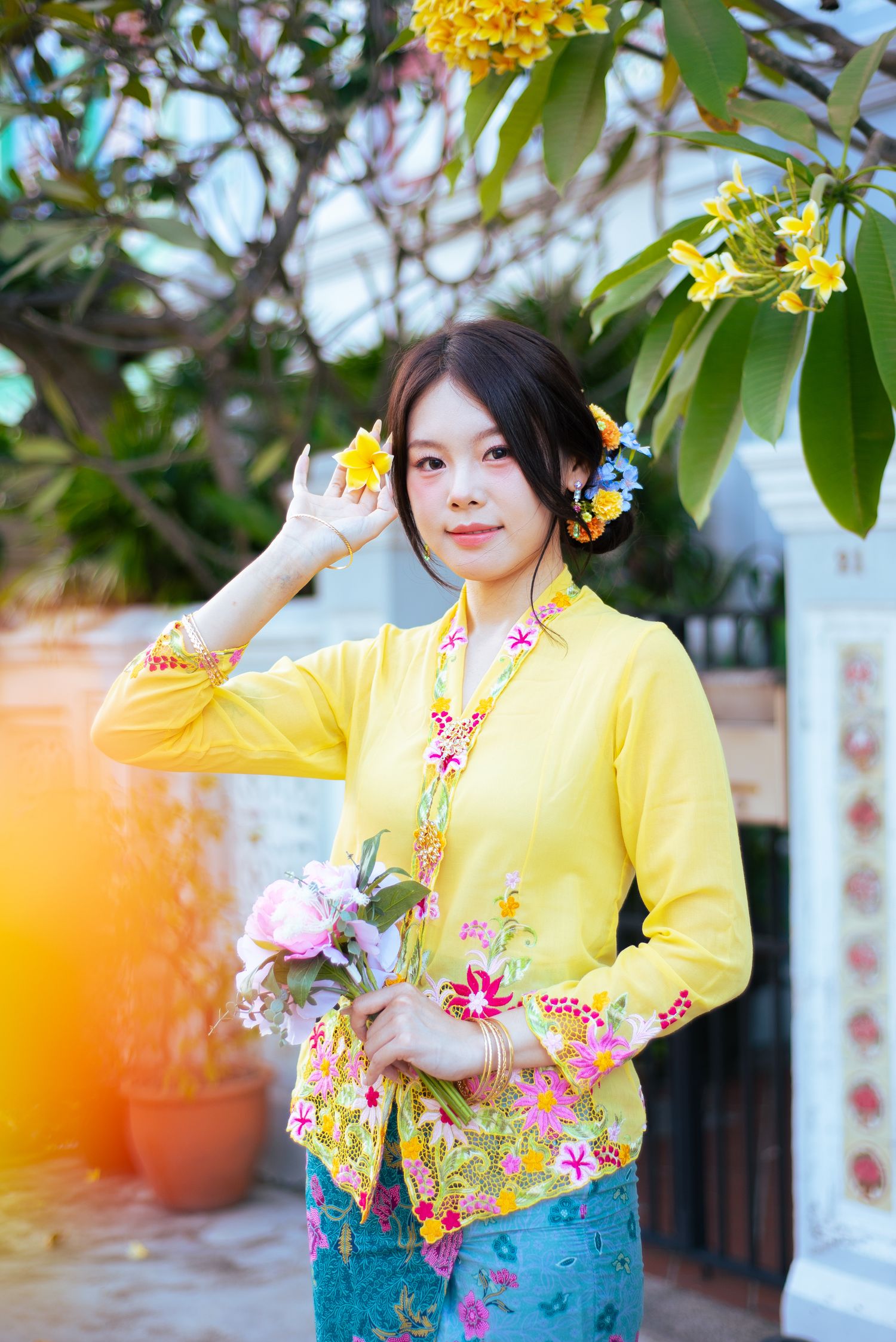 A person in a bright yellow floral kebaya top and blue skirt poses outdoors with tropical flowers and foliage in the background.