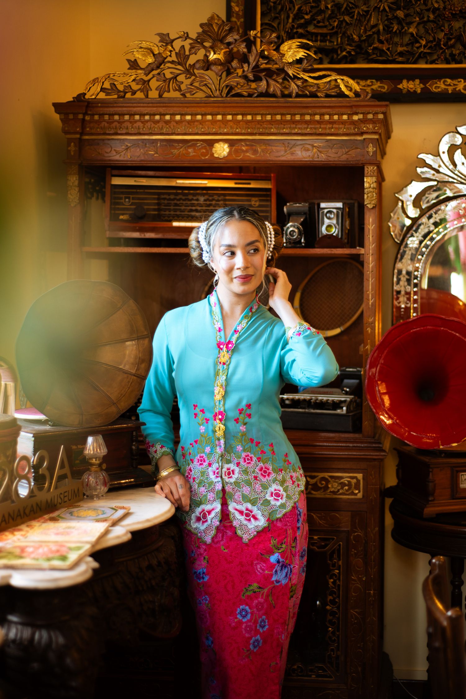 A vintage room interior with an ornate wooden cabinet, phonograph, and figure wearing turquoise and pink floral clothing.