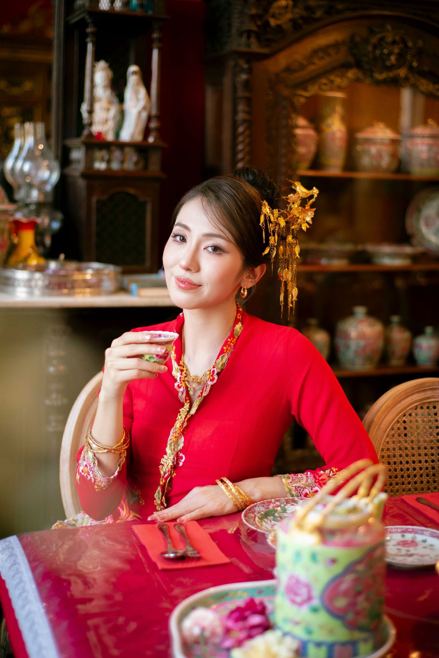 Elegant woman in traditional red dress with gold accessories enjoys tea service at ornate table with antique decor.