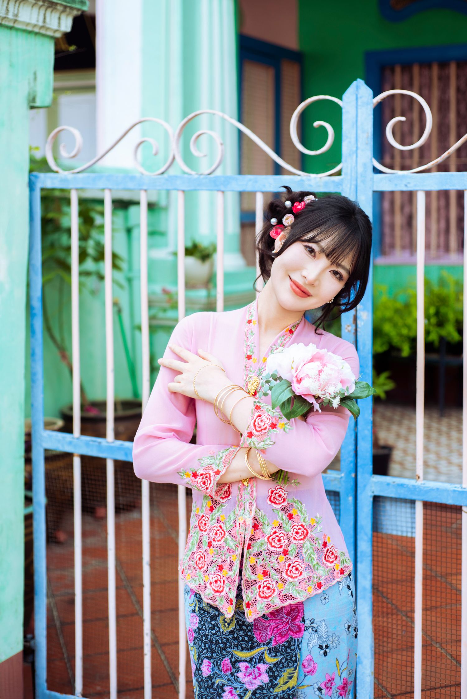 A feminine figure in a pink floral top and patterned skirt poses gracefully by a turquoise gate with flowers.