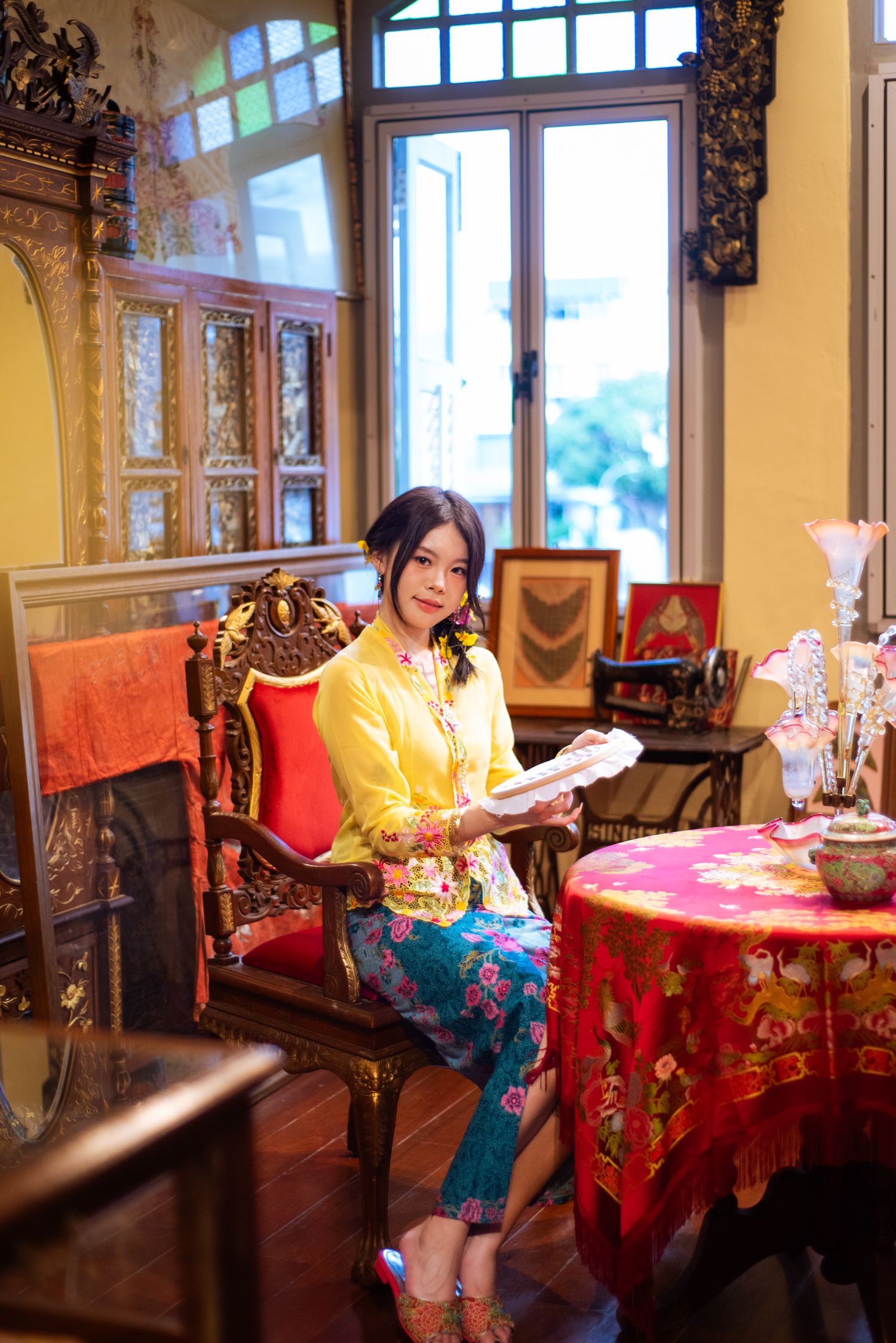 Woman in yellow traditional Asian attire reads while seated in an ornate vintage parlor with red accents and decorative details.