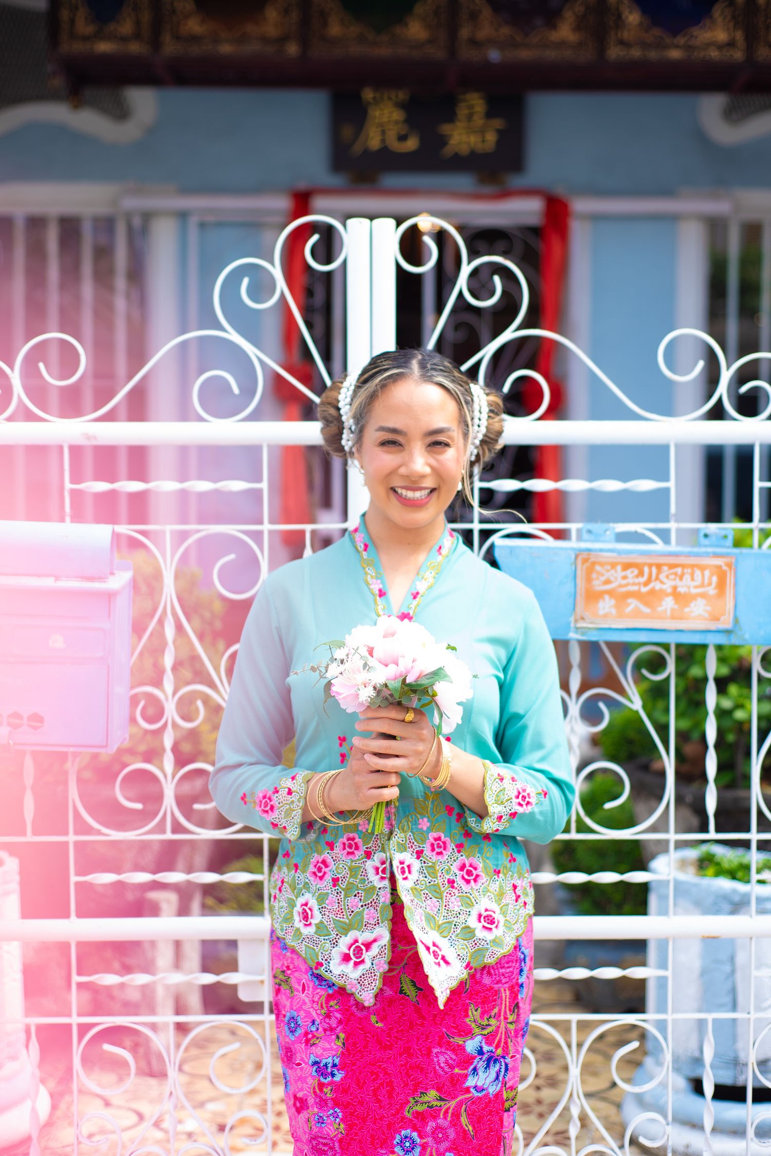 Smiling figure in traditional floral Asian clothing stands by ornate white gate holding pink flowers.