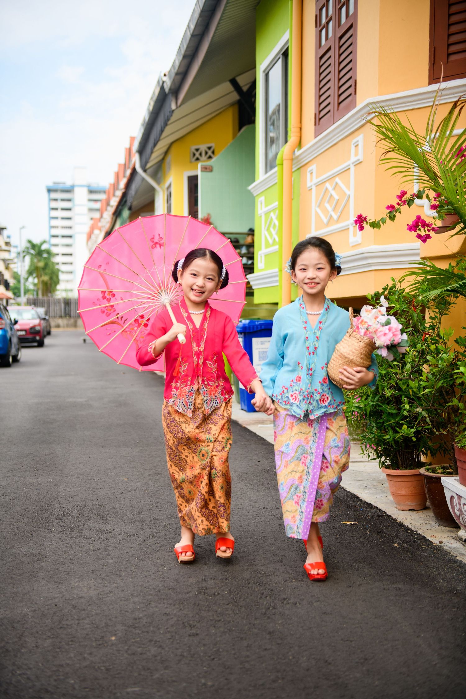 Two young girls in colorful traditional Peranakan kebaya outfits walk down a vibrant street, one holding a pink parasol.