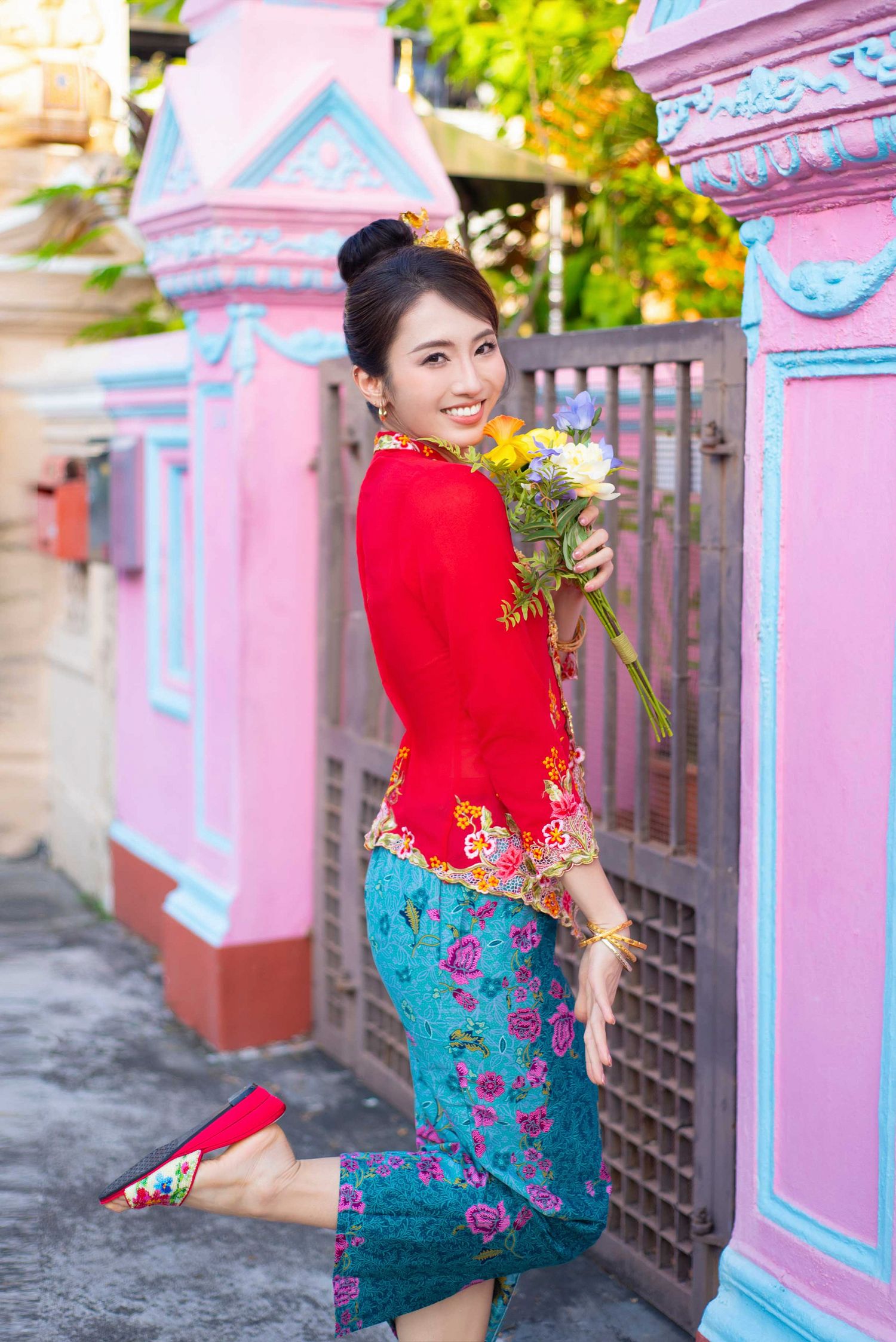 Person in traditional attire with red top and floral turquoise skirt poses by pink architectural details holding flowers.